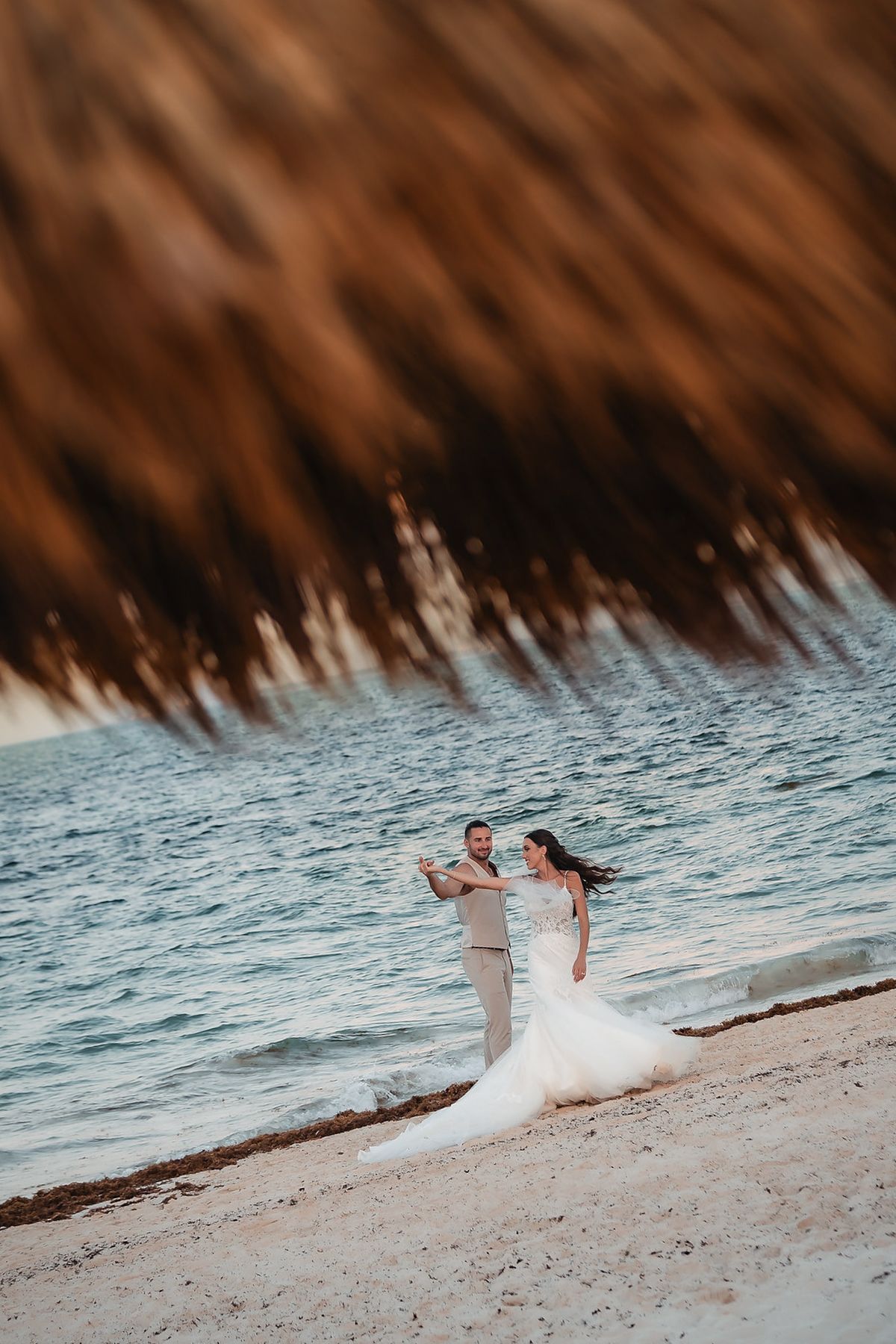 Bride and groom dancing on a sandy beach at sunset with ocean waves behind them during a romantic destination wedding.