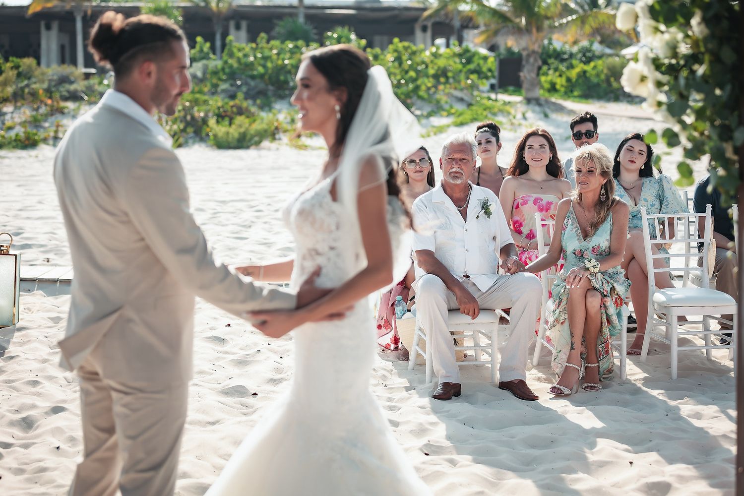 Bride and groom hold hands during an outdoor beach wedding ceremony with guests seated on white chairs in the background.