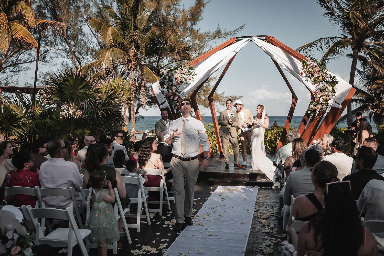 Outdoor beach wedding ceremony with guests seated on white chairs, couple at floral arch altar, palm trees and ocean backdrop.