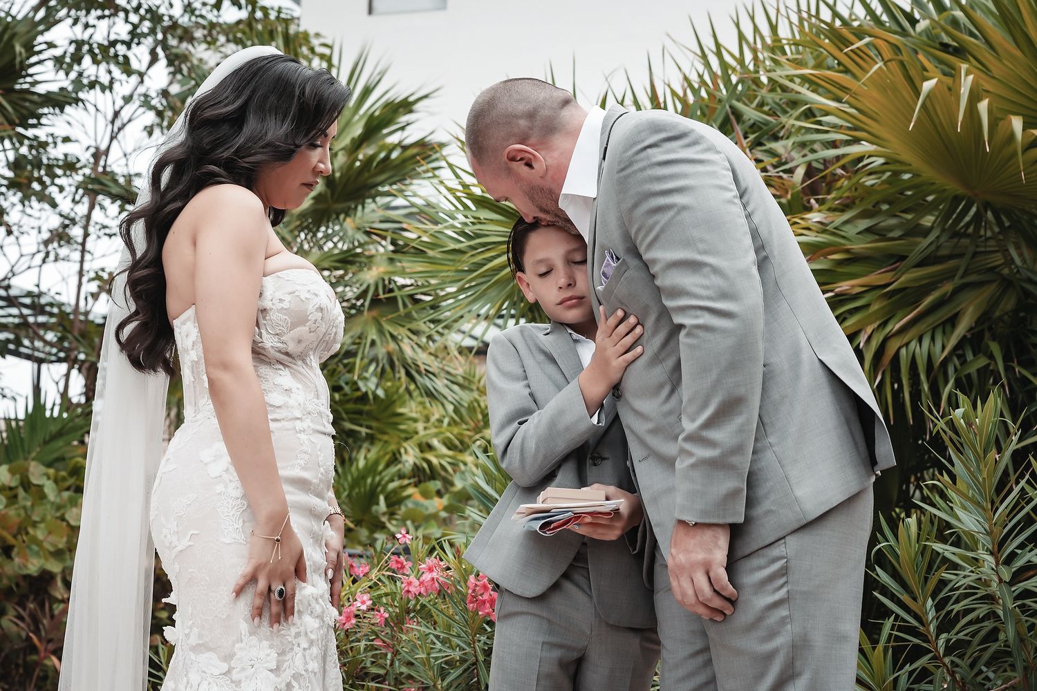 Bride and groom in gray suit share tender moment with young boy during outdoor wedding ceremony surrounded by tropical plants.