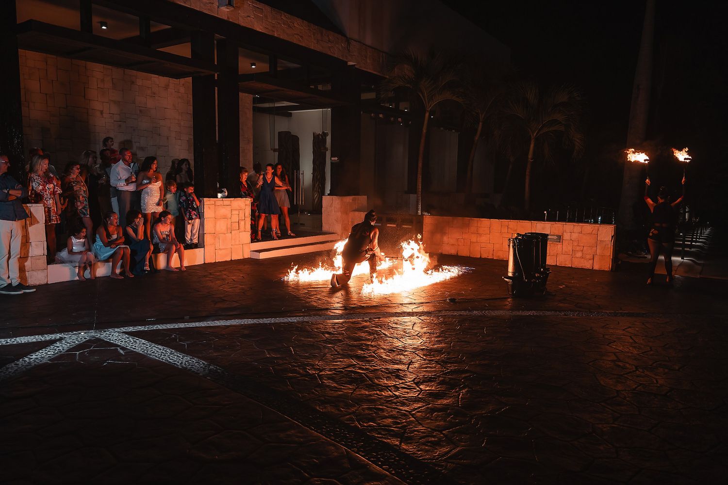 A fire performer dances with flames at night while spectators watch from the sidelines at an outdoor event.