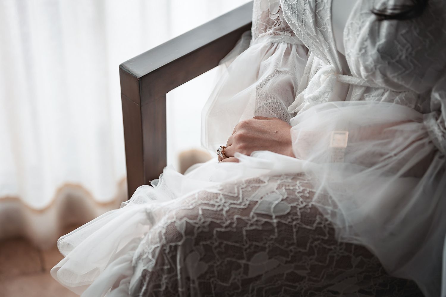 Bride in white lace wedding dress sitting gracefully in a wooden chair near a sunlit window.