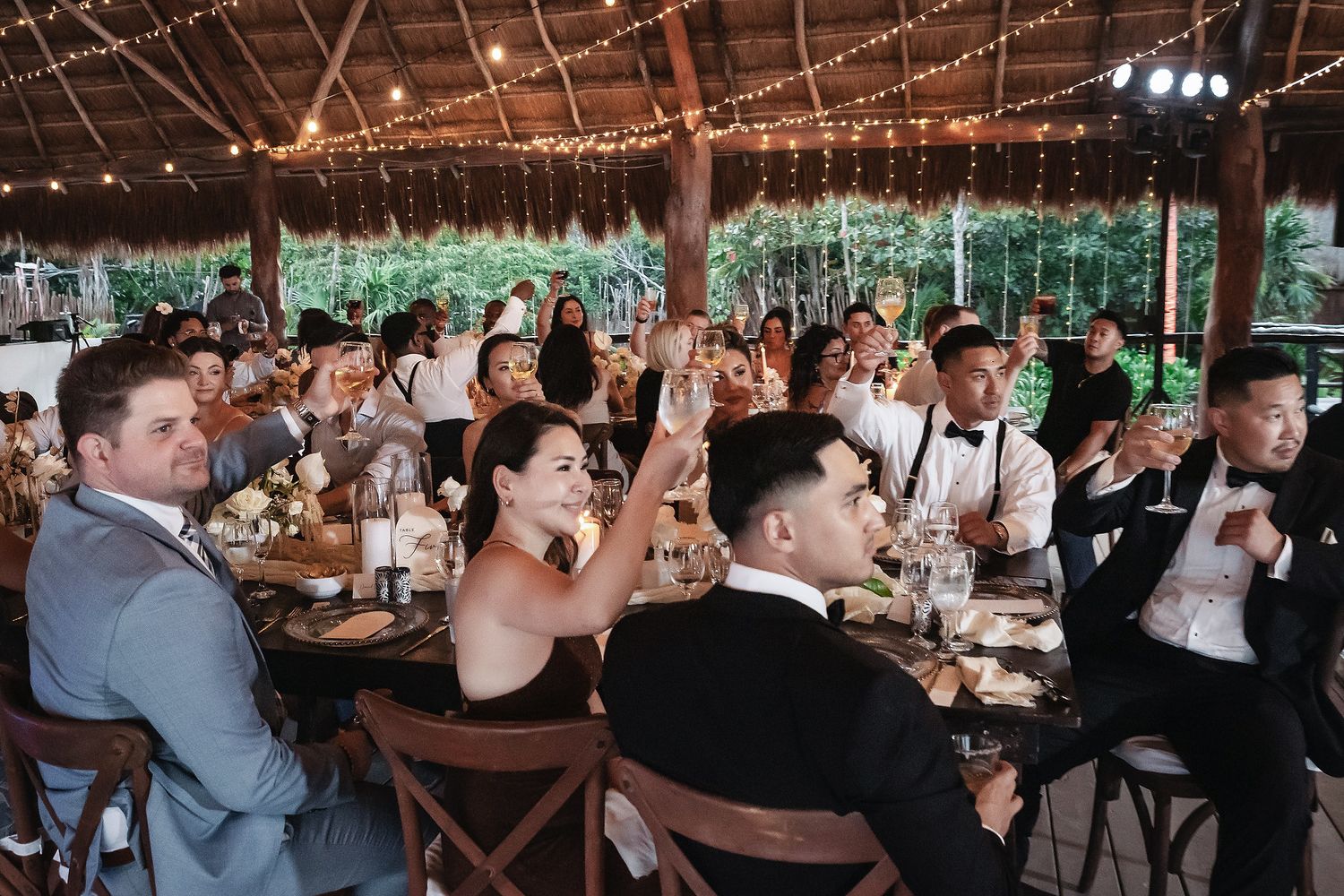 Guests raising glasses in a toast at an elegant outdoor wedding reception under string lights and a wooden pergola.