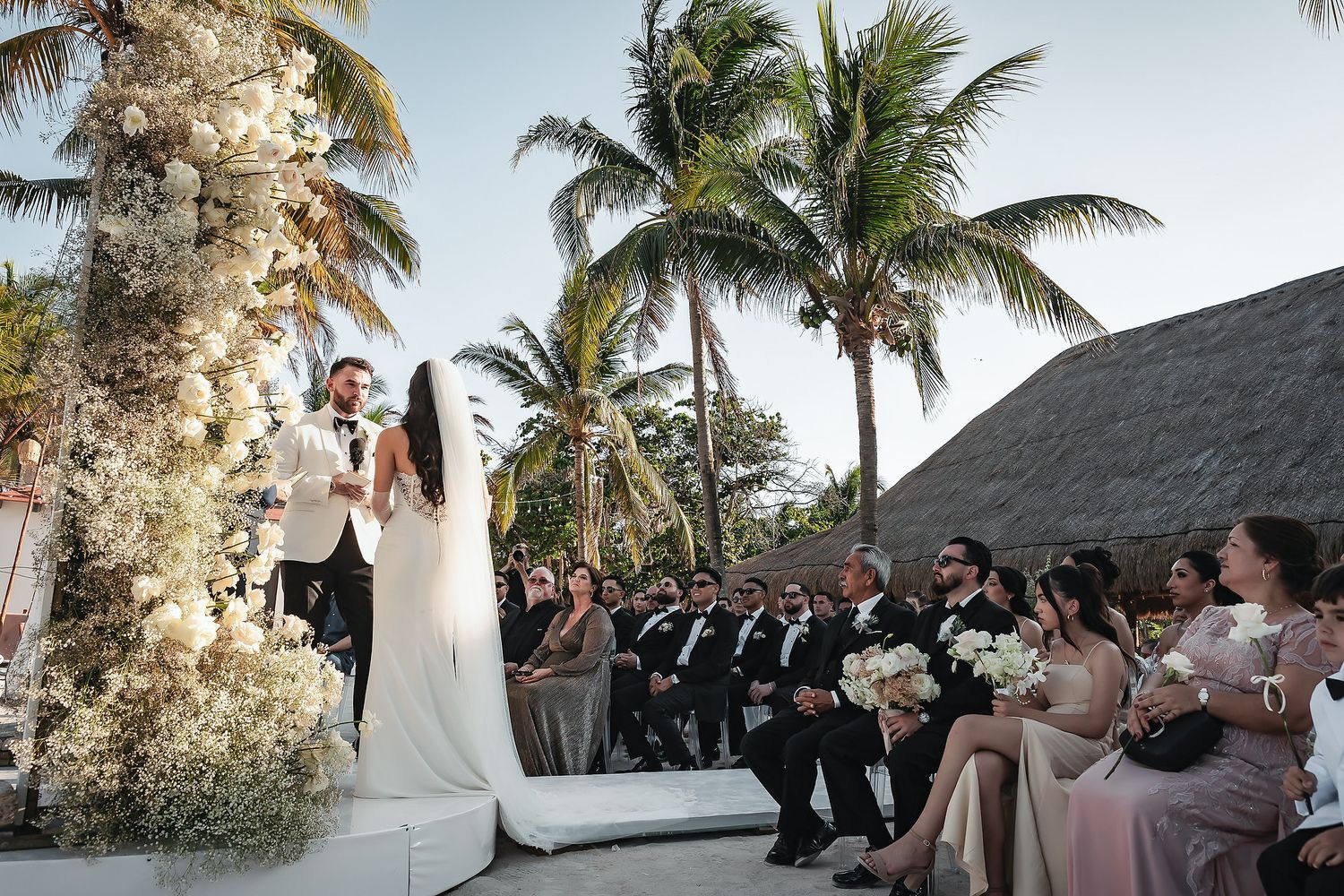 Outdoor beach wedding ceremony with bride and groom at floral arch, palm trees, and guests seated in tropical venue.