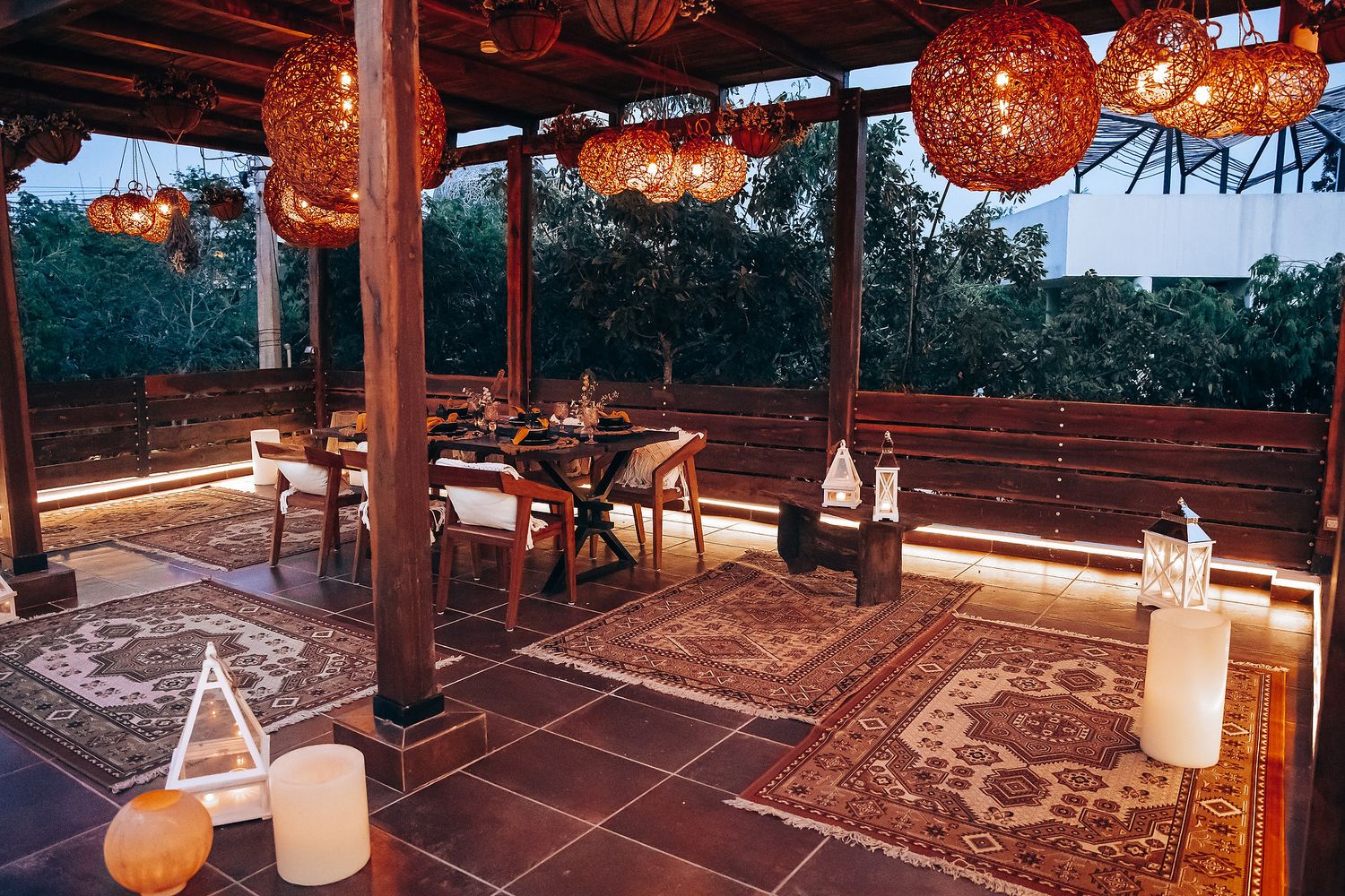 Bohemian outdoor evening dining setup with woven pendant lights, patterned rugs, and wooden pergola at dusk.