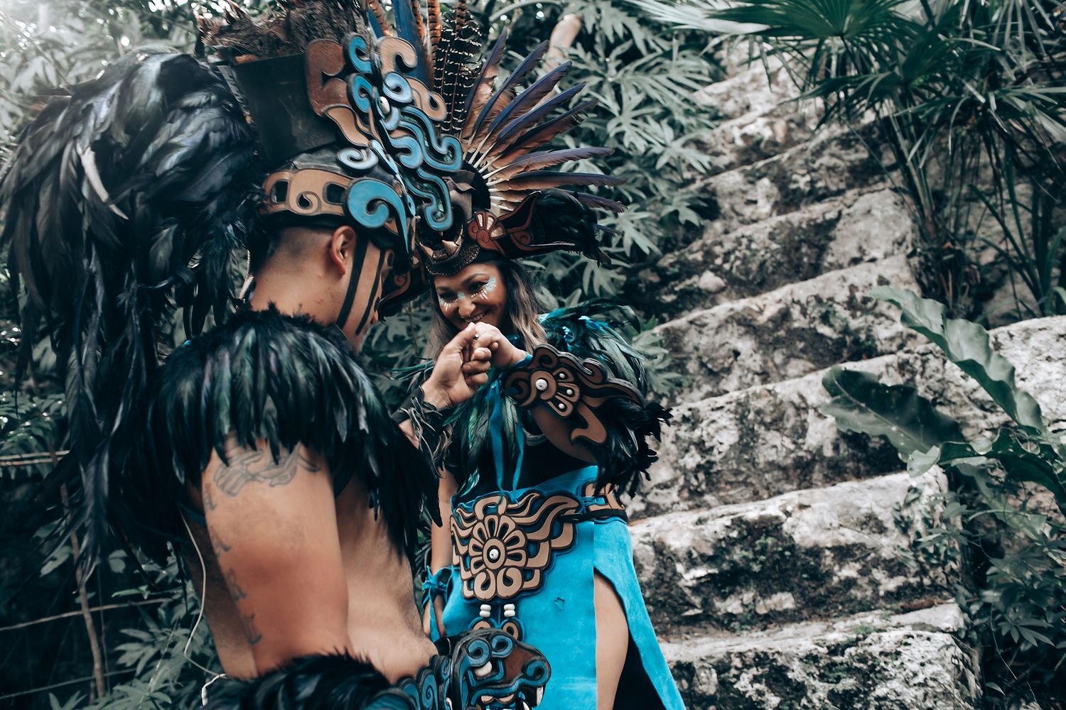 Two performers dressed in elaborate Mayan ceremonial costumes with turquoise and feathered headdresses in a jungle setting.