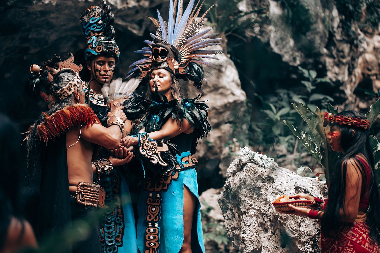 Group of performers in elaborate Aztec ceremonial costumes gather together near rocky cave walls.