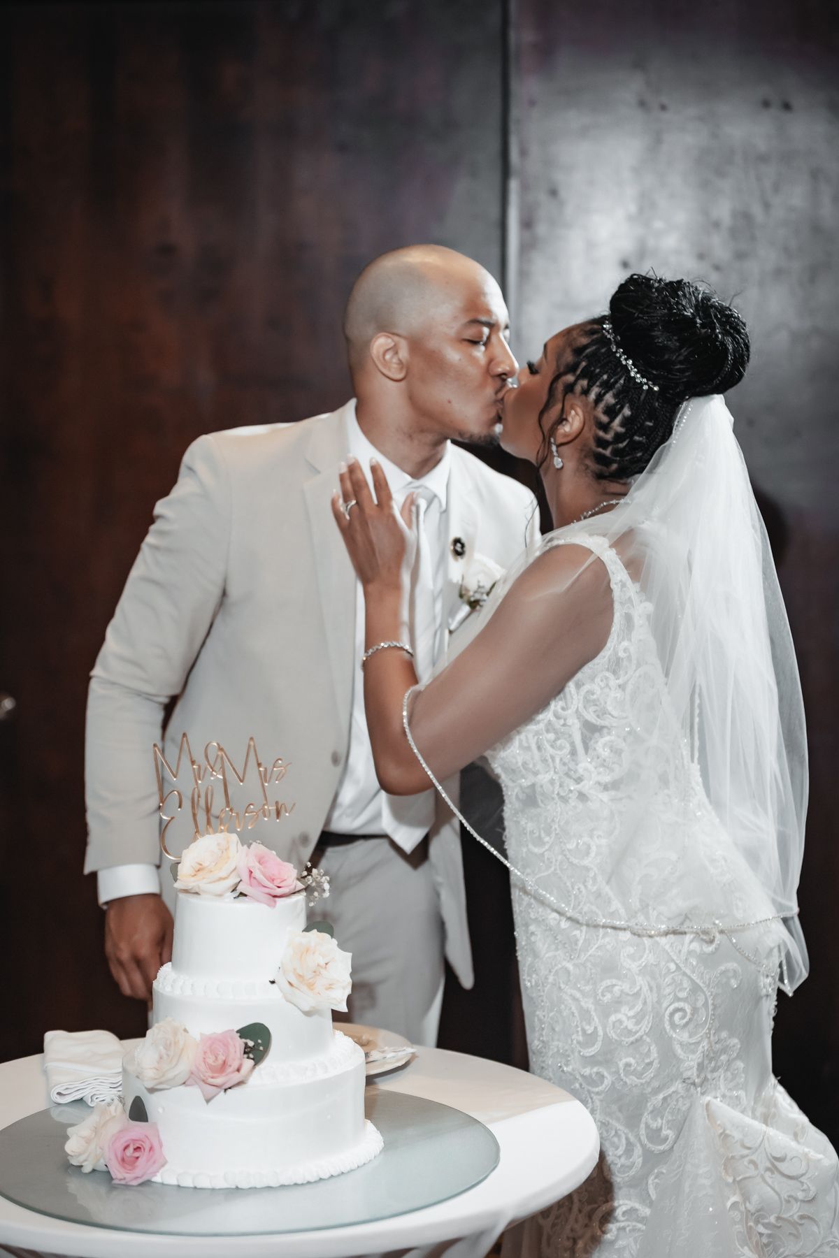 Newlywed couple sharing a kiss beside a white floral wedding cake during their elegant indoor reception ceremony.