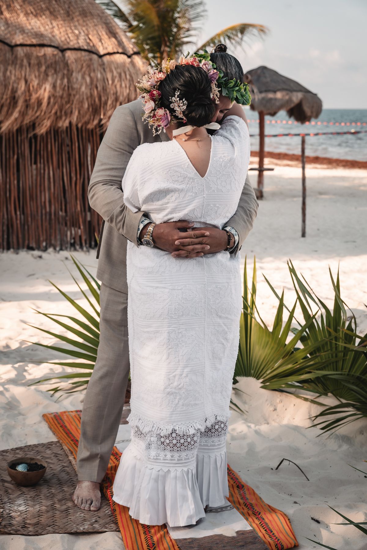 Bride in white lace dress with floral crown embraced by groom on sandy beach near thatched huts at sunset.