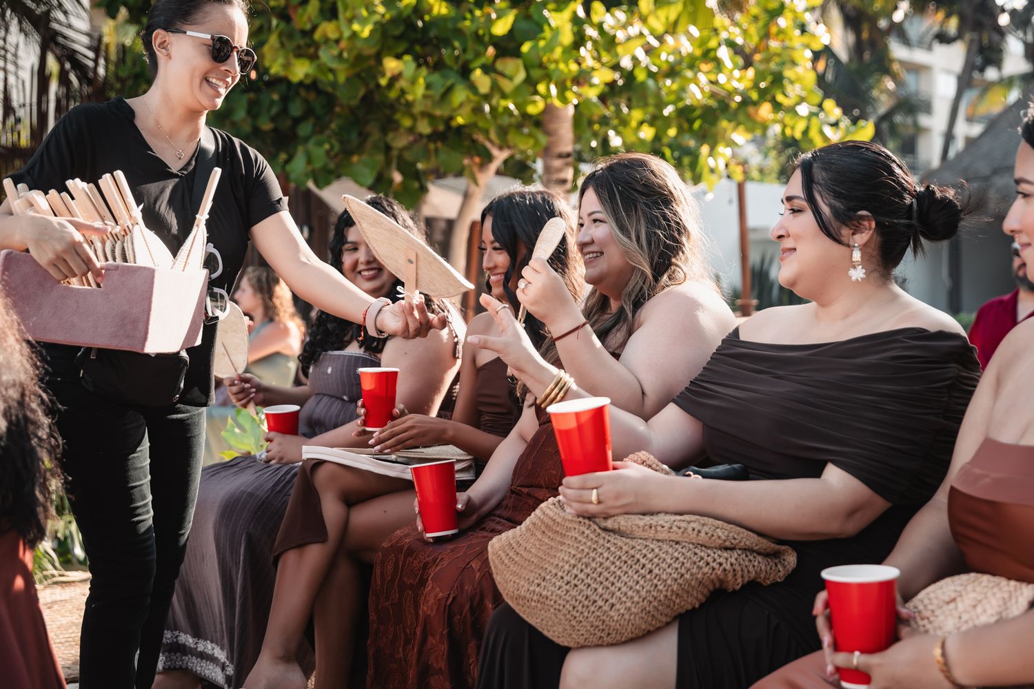 Women sitting outdoors at a party, sharing food and red solo cups, smiling and socializing in warm sunlight.