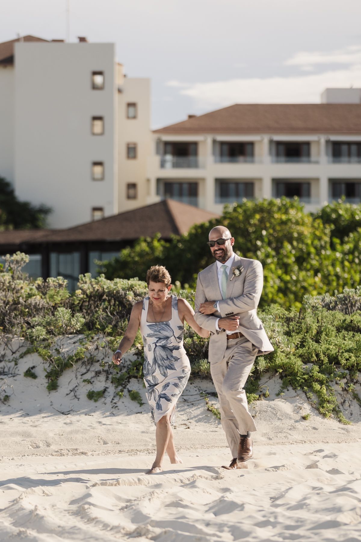 Couple in wedding attire walking hand-in-hand on sandy beach with lush greenery and resort building in background.