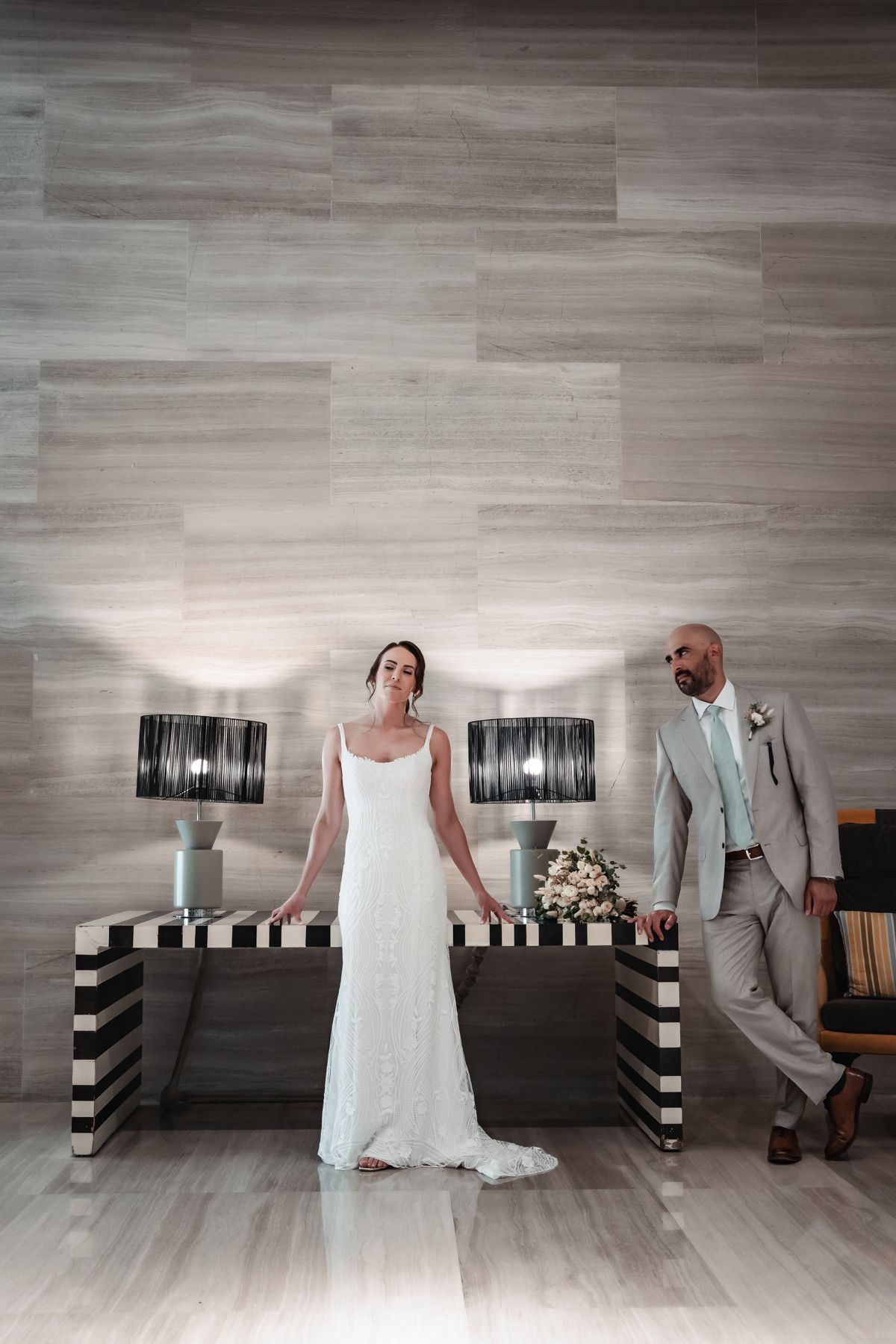 Bride in white gown and groom in gray suit pose elegantly beside a striped console table with black lamps.