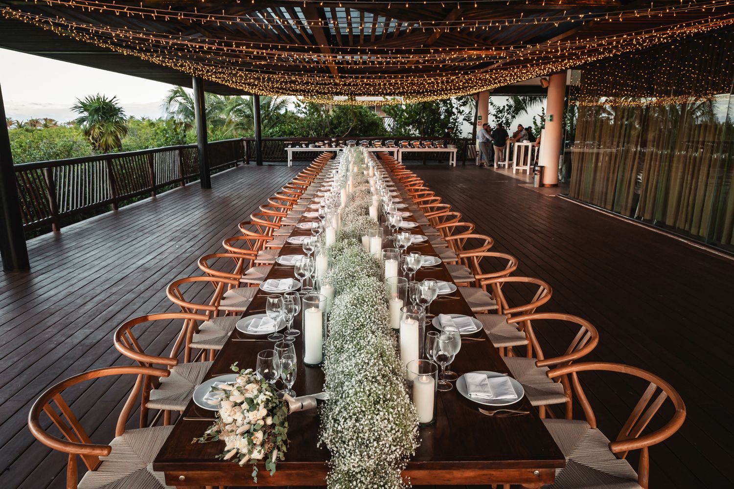 Elegant outdoor wedding reception on wooden deck with long tables, fairy lights, floral runners, and candles at dusk.