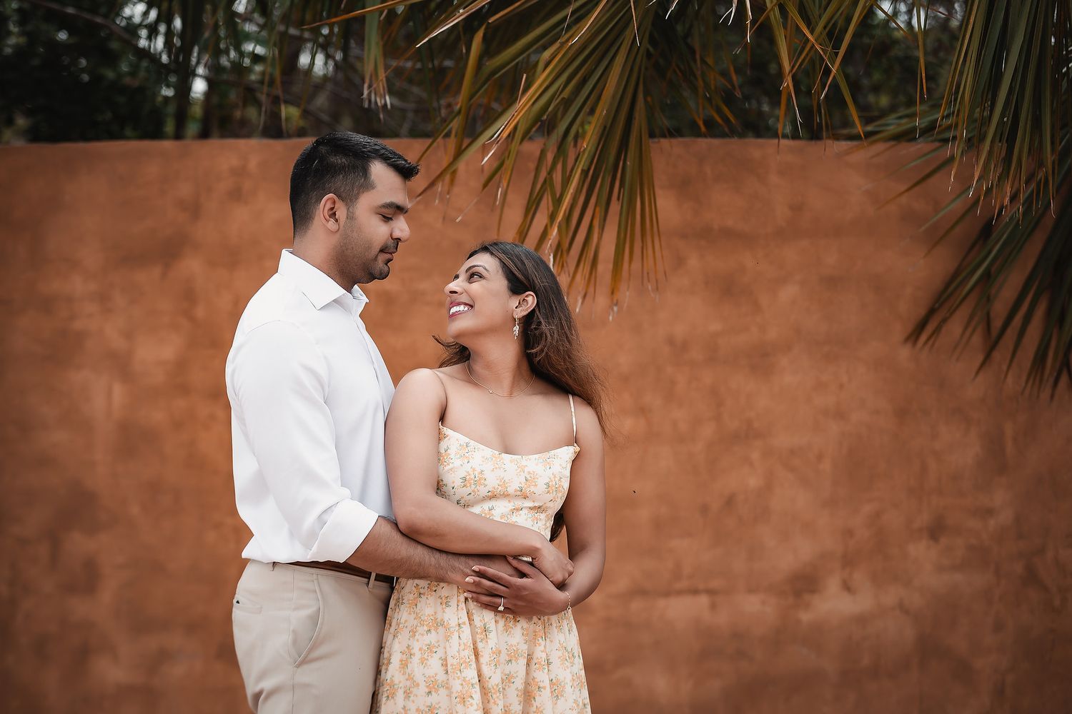 Couple smiling at each other in front of a terracotta wall with palm leaves, wearing casual elegant attire.