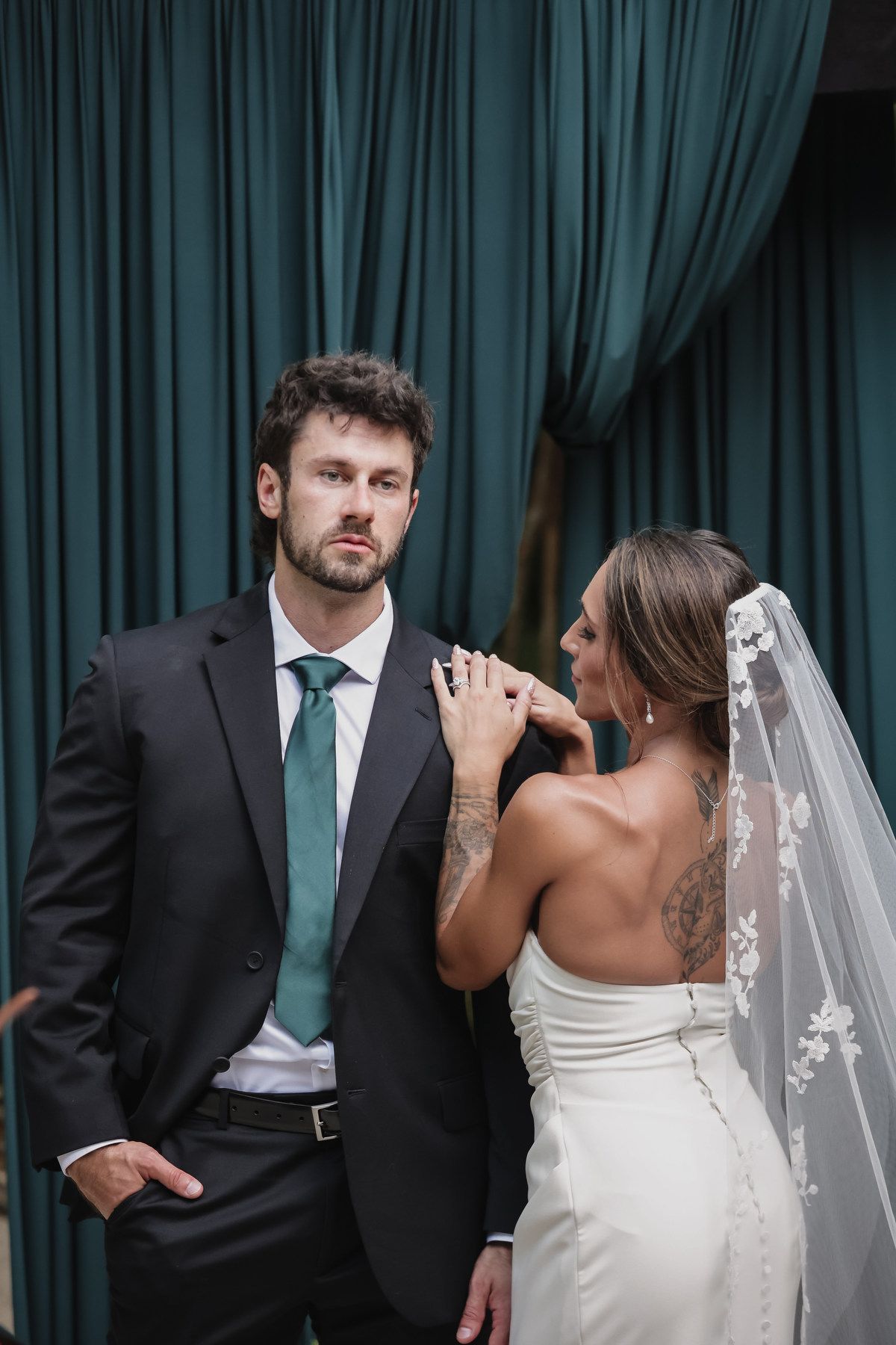 Bride in white gown and veil with groom in dark suit and teal tie posing before green curtains.