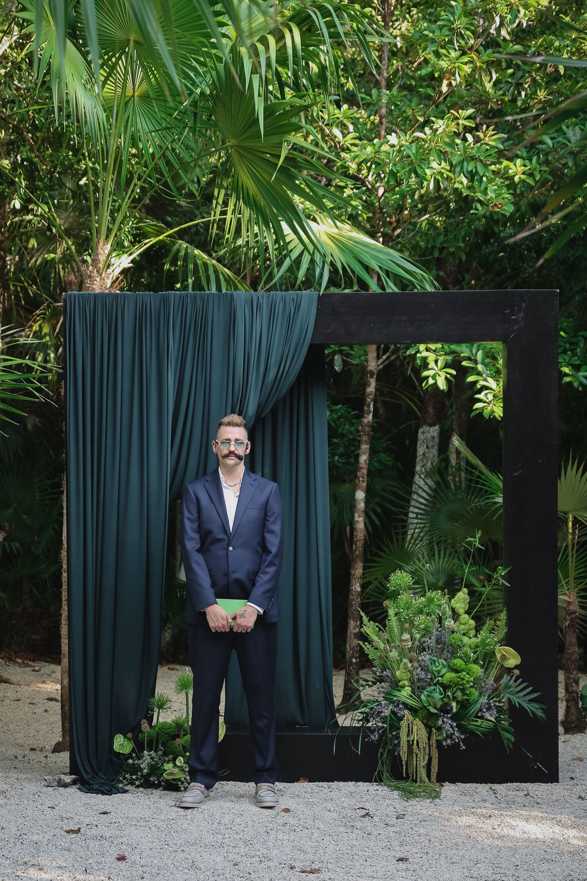 Bearded groom in navy suit stands before black frame altar with emerald green draping and tropical greenery outdoors.