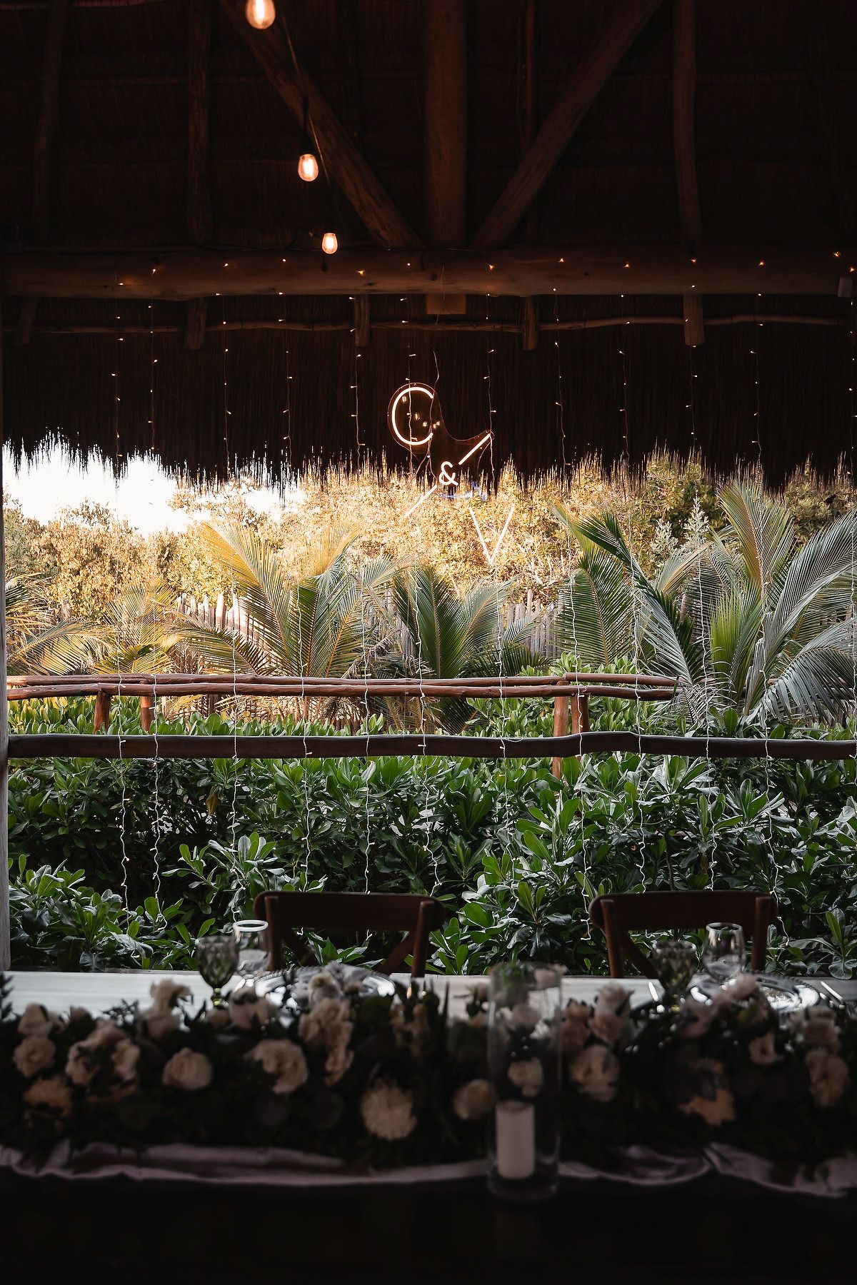Elegant tropical wedding reception venue with fairy lights, palm trees, floral table decor, and a glowing monogram backdrop.
