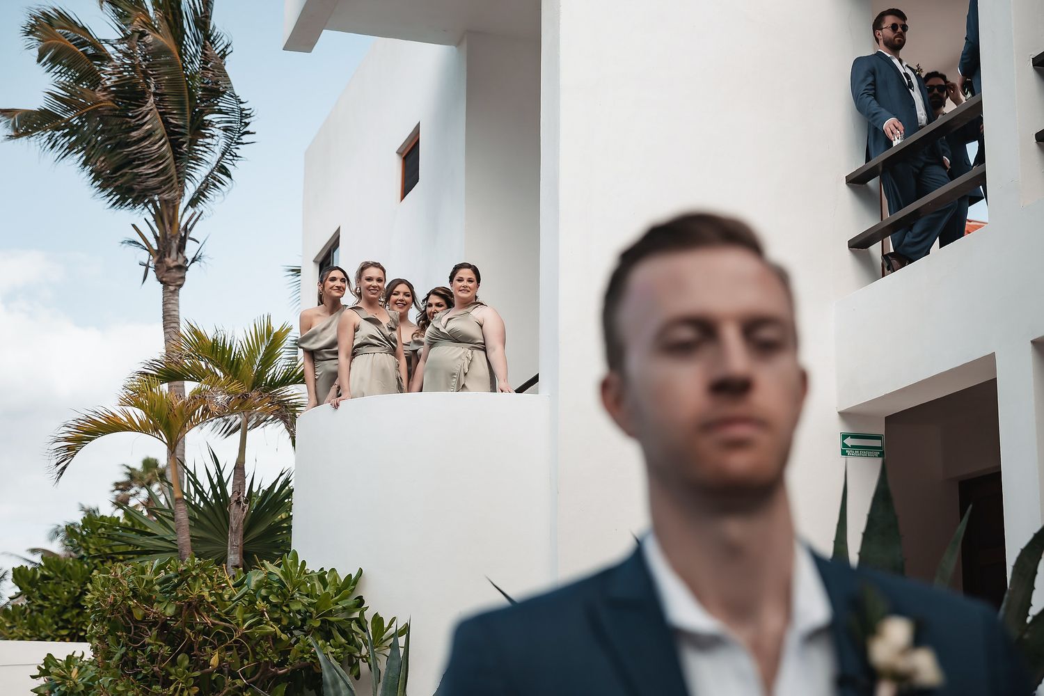 Groom stands in foreground while bridesmaids watch from a white balcony at a tropical wedding venue.