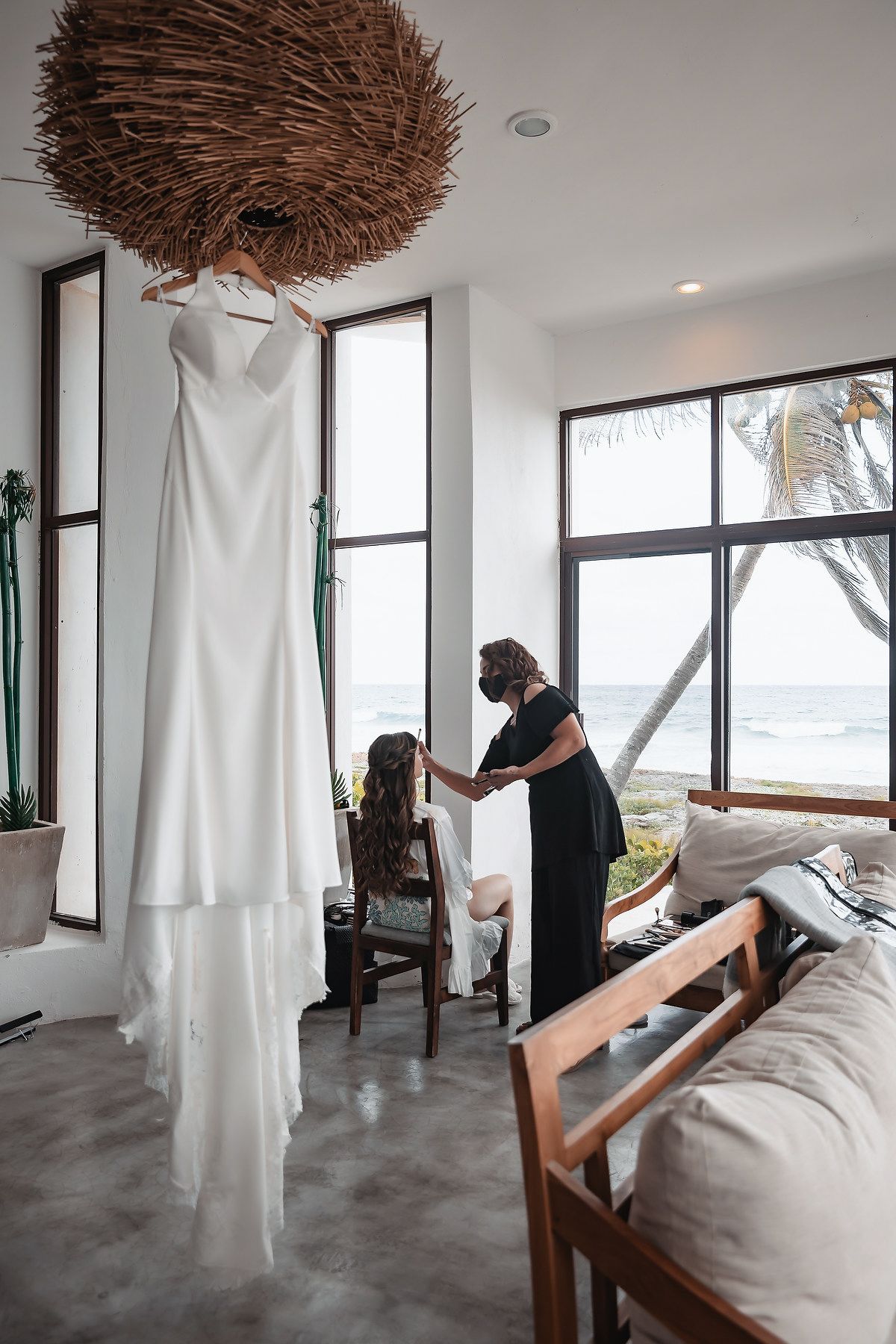 White wedding dress hanging near ocean-view windows as stylist prepares bride in a modern beachfront room.