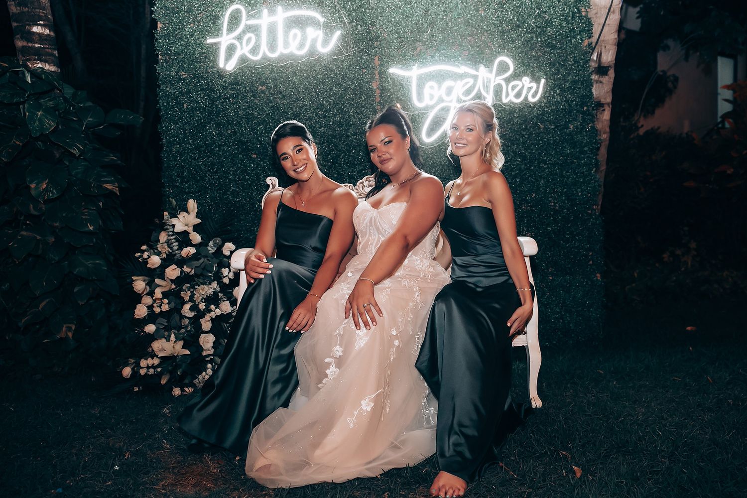 Three women in formal attire pose against a dark wall with neon signage and floral decorations.