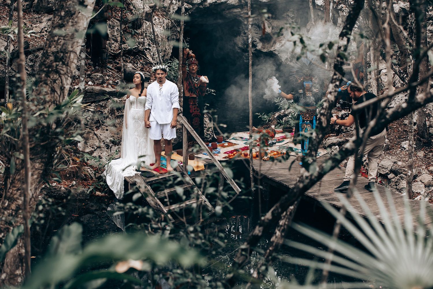 Couple in white outfits pose together in a tropical garden setting with moody natural lighting and foliage.
