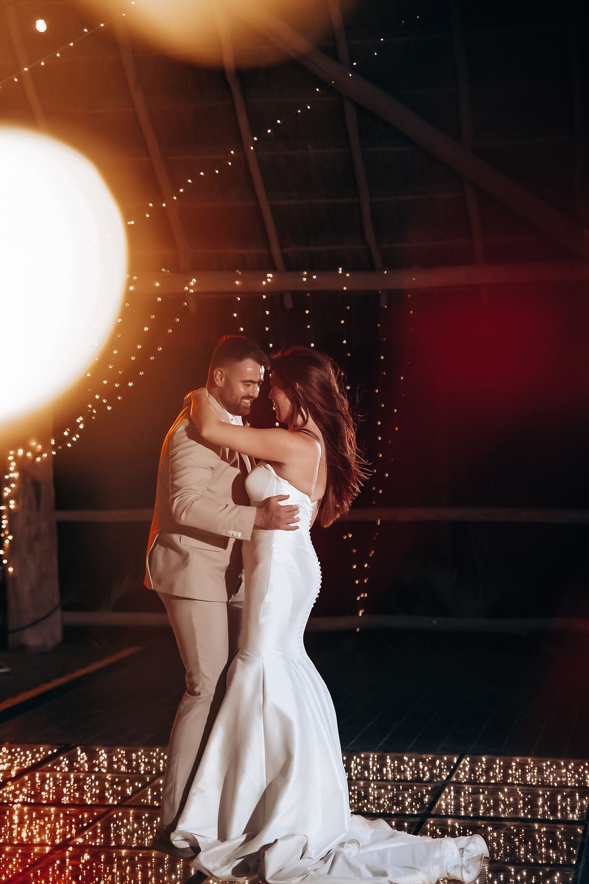 Romantic couple dances beneath glowing string lights at an evening wedding reception.
