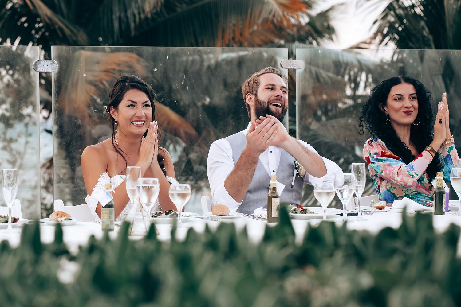Wedding reception table settings with glassware and tropical decor under palm trees.