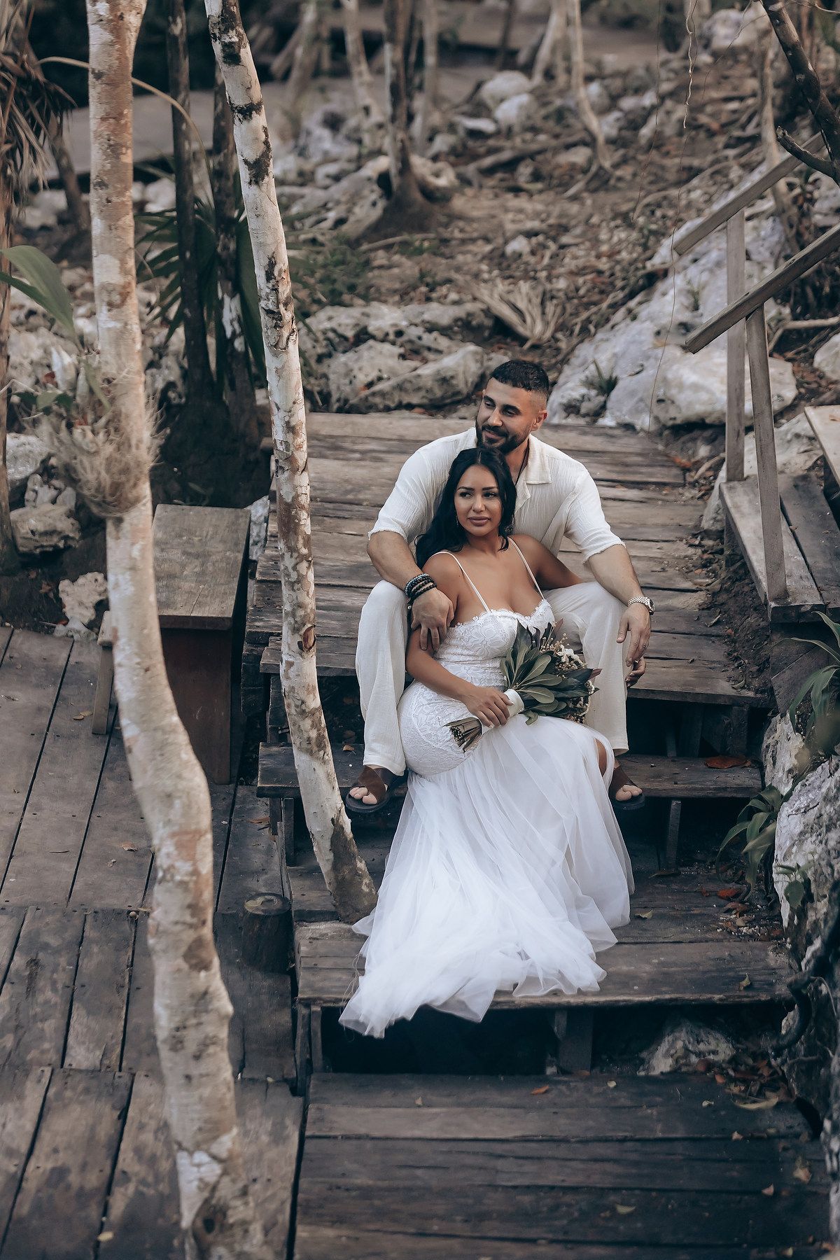 A couple poses on wooden steps in a forest setting wearing elegant white wedding attire.