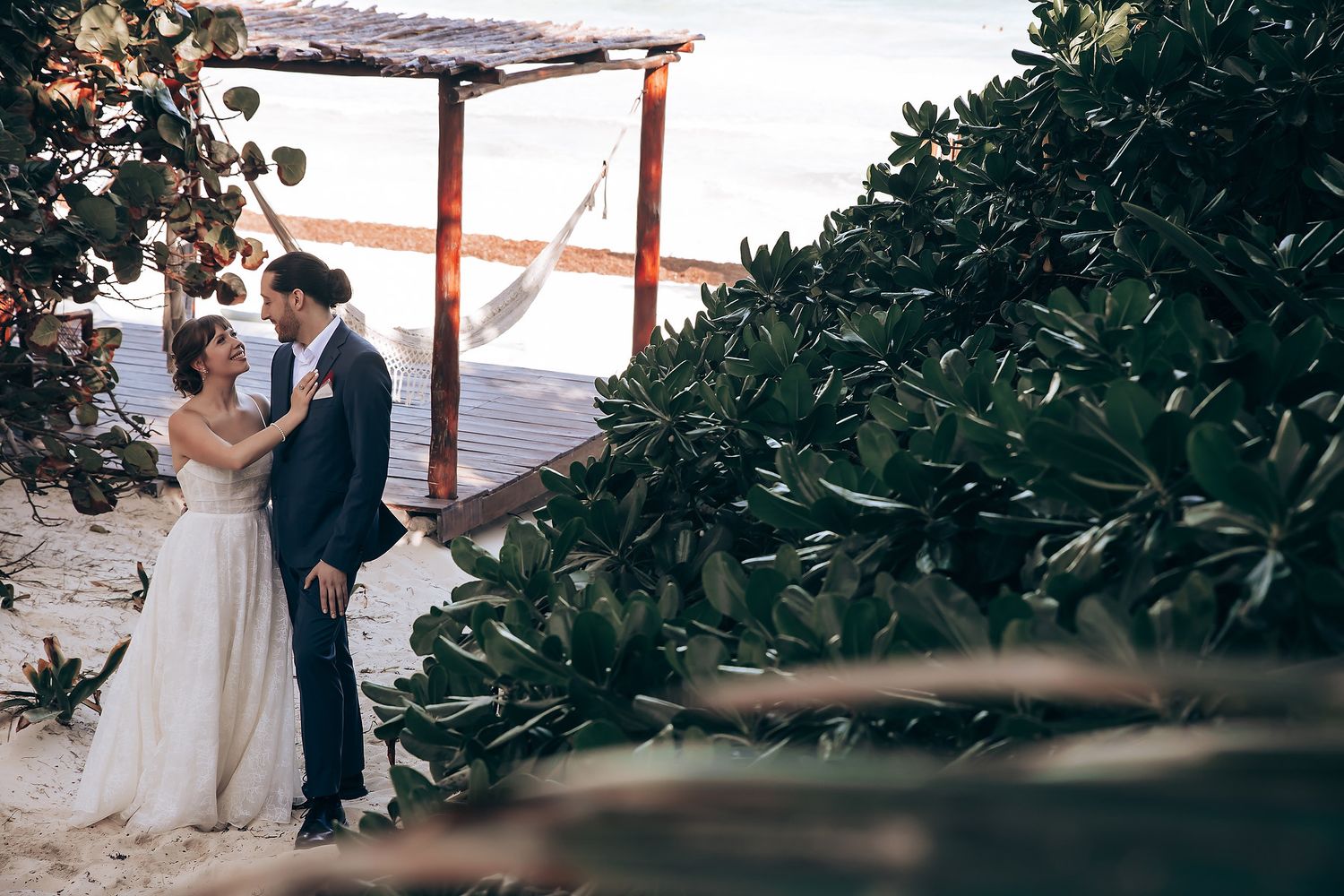 A couple stands together during an outdoor wedding ceremony beneath a rustic wooden pergola with greenery.