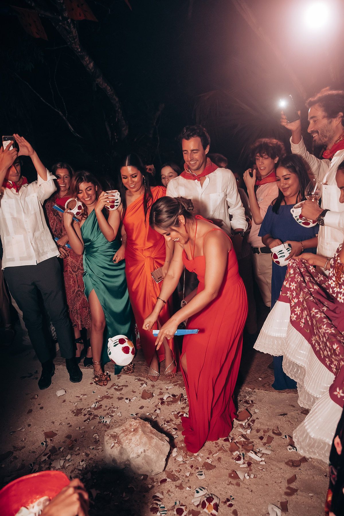 Party guests in colorful dresses dance and celebrate on a sandy beach at night.