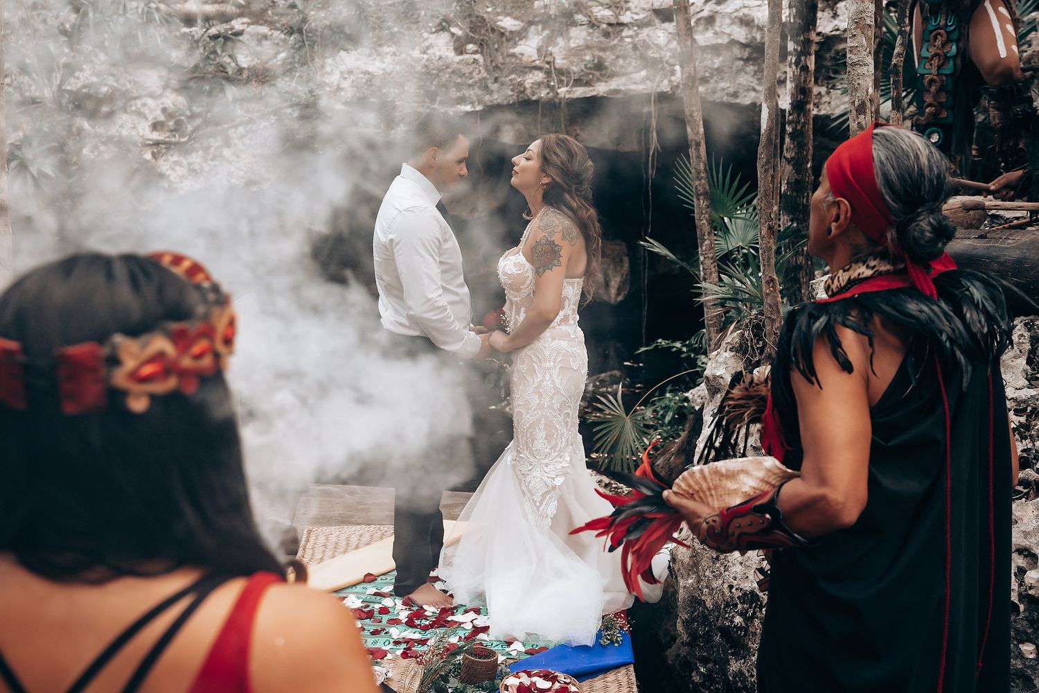 A couple participates in a mystical outdoor wedding ceremony surrounded by smoke and ceremony attendees in traditional dress.