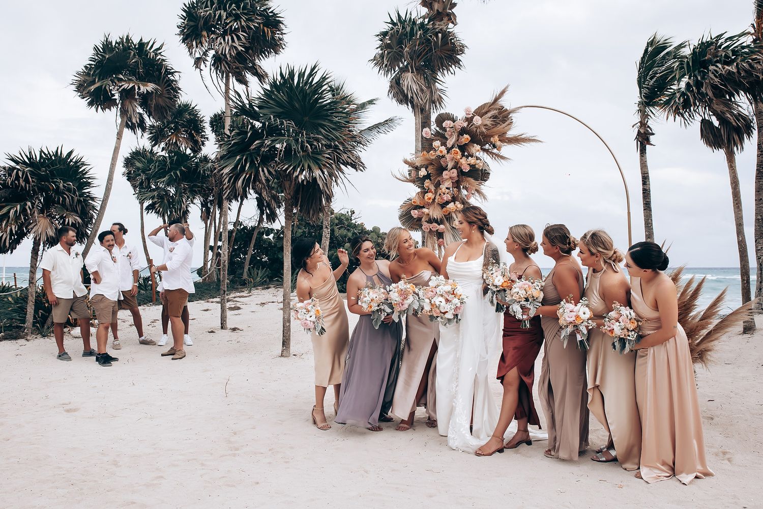 Bridal party in coordinated neutral dresses poses along a palm-lined beach.