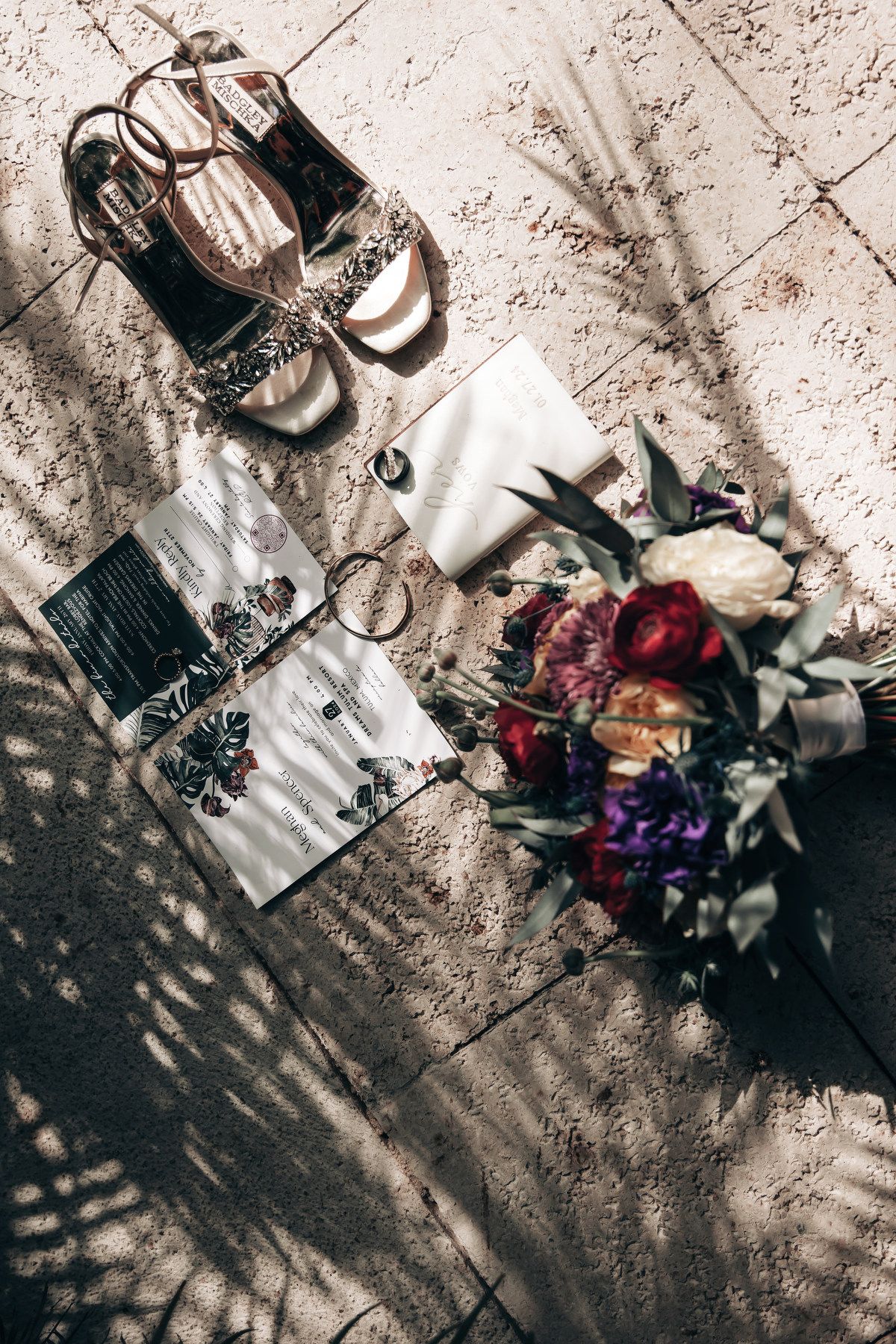 Bridal flat lay featuring heels, bouquet, rings, clutch, and invitations on textured stone floor in natural light.