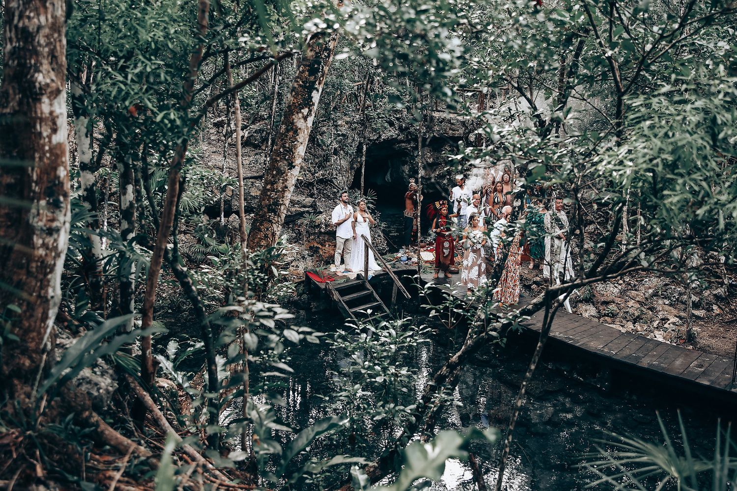 Guests in white clothing stand together in a wooded outdoor ceremony space.