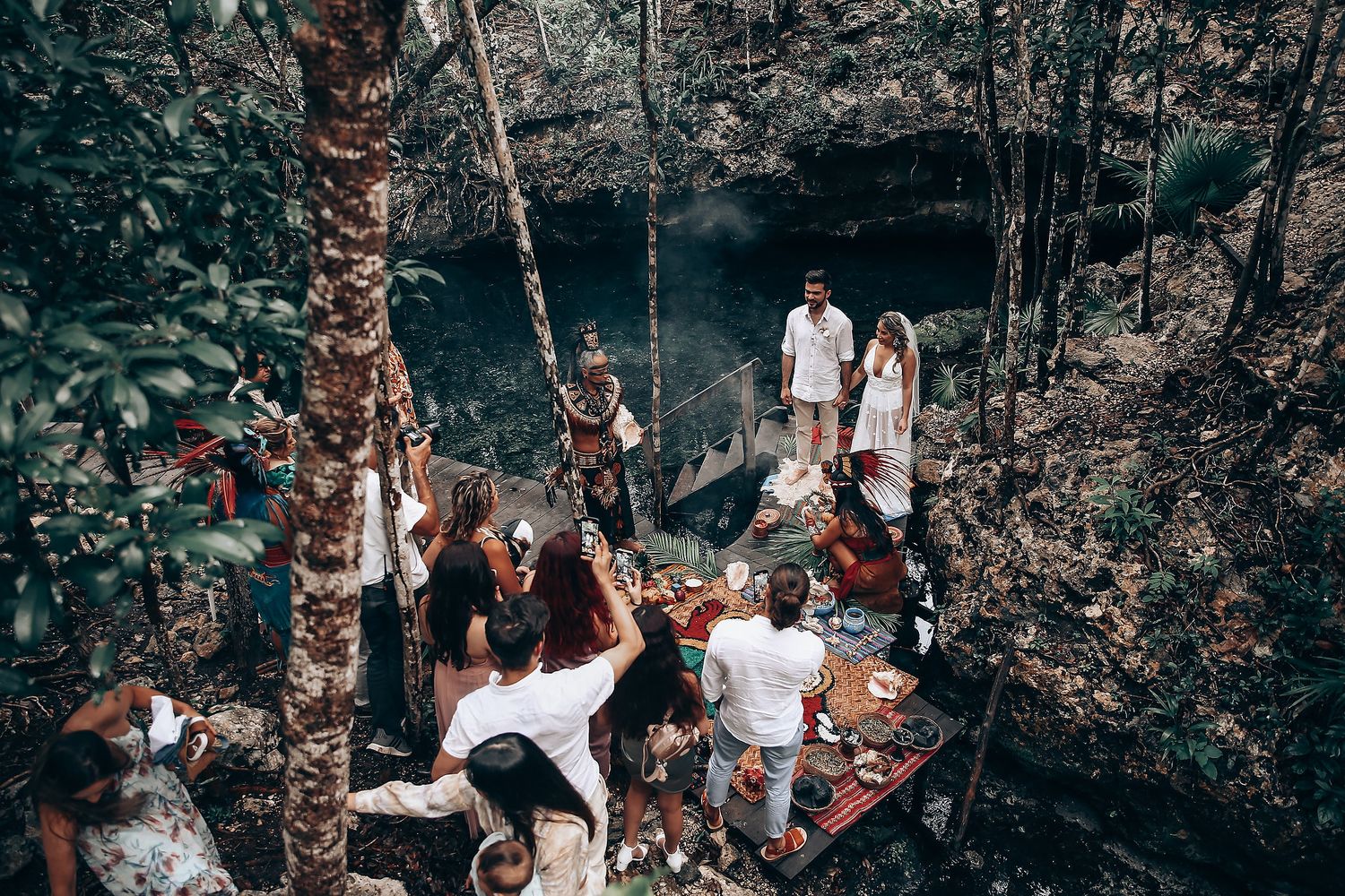 Wedding guests gather in a lush forest setting wearing white attire for an outdoor celebration.