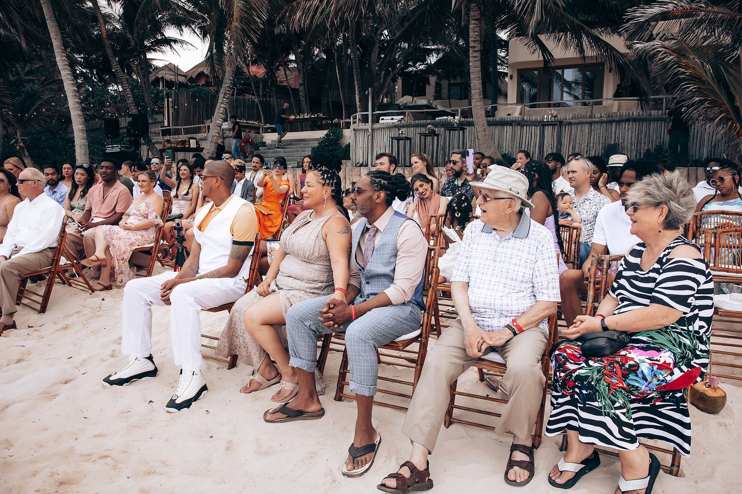 Wedding guests seated in casual attire watching beachside ceremony.