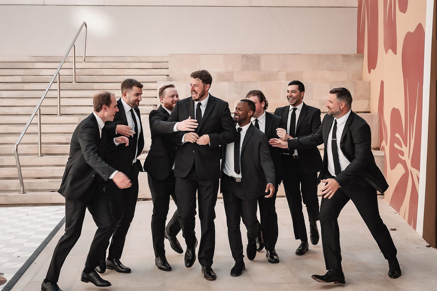 Groomsmen in black suits pose playfully in a line against a light colored wall.