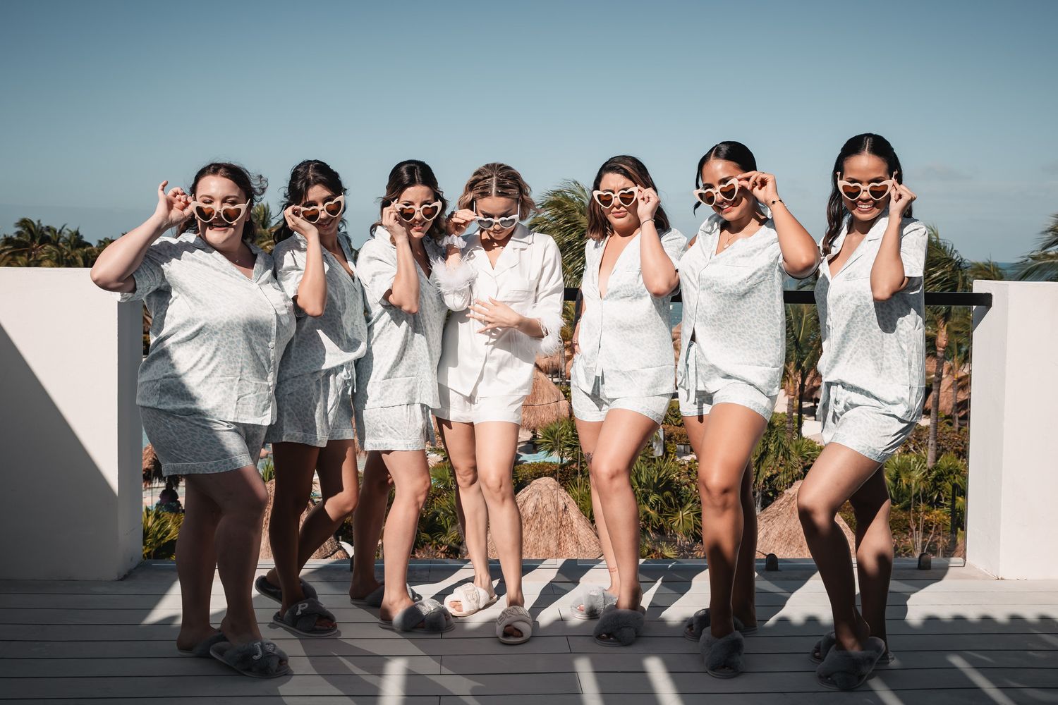 Group of bridesmaids in matching white outfits posing with sunglasses on a sunny rooftop.