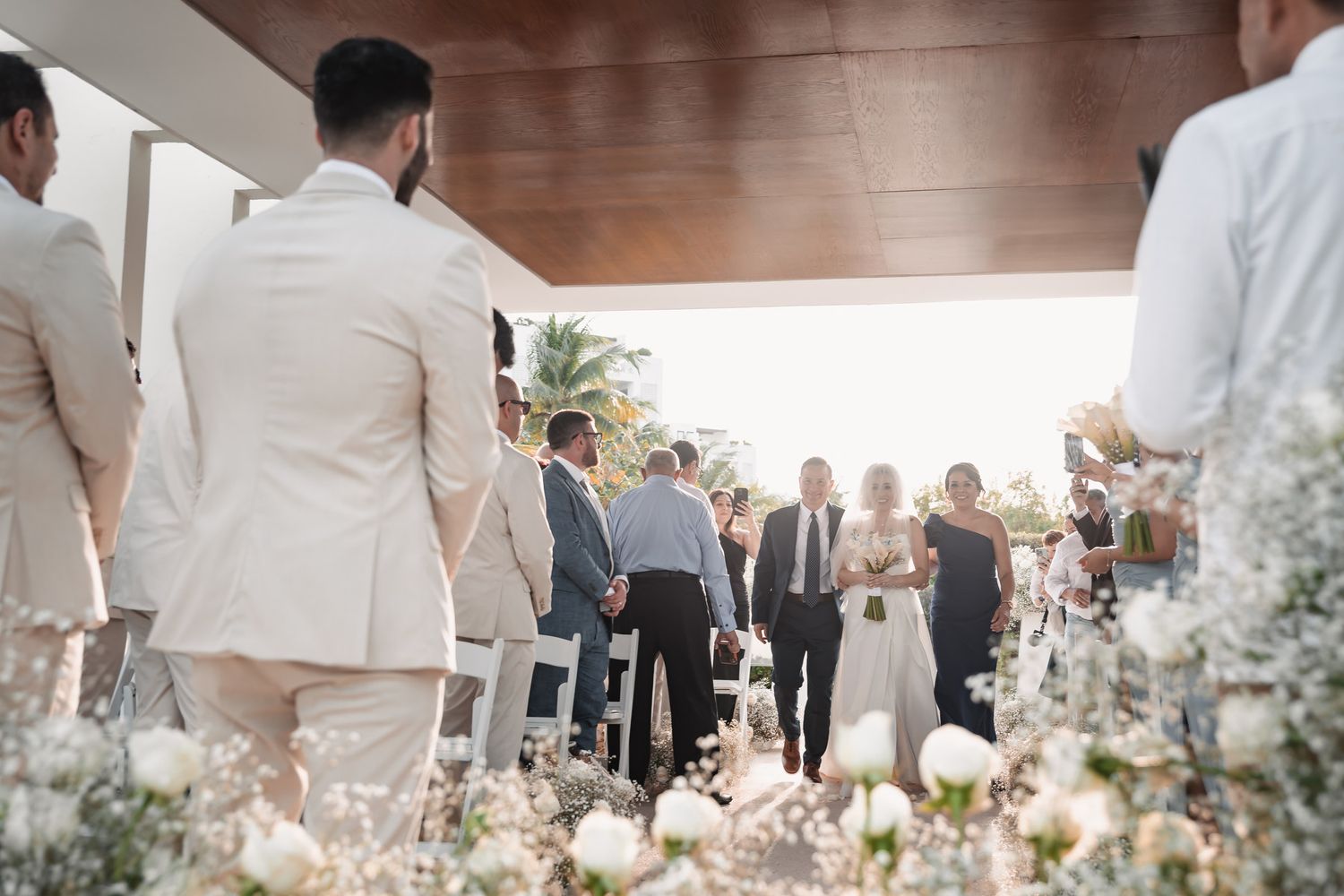 Guests gather as bride walks down the aisle at a sunlit outdoor wedding ceremony.