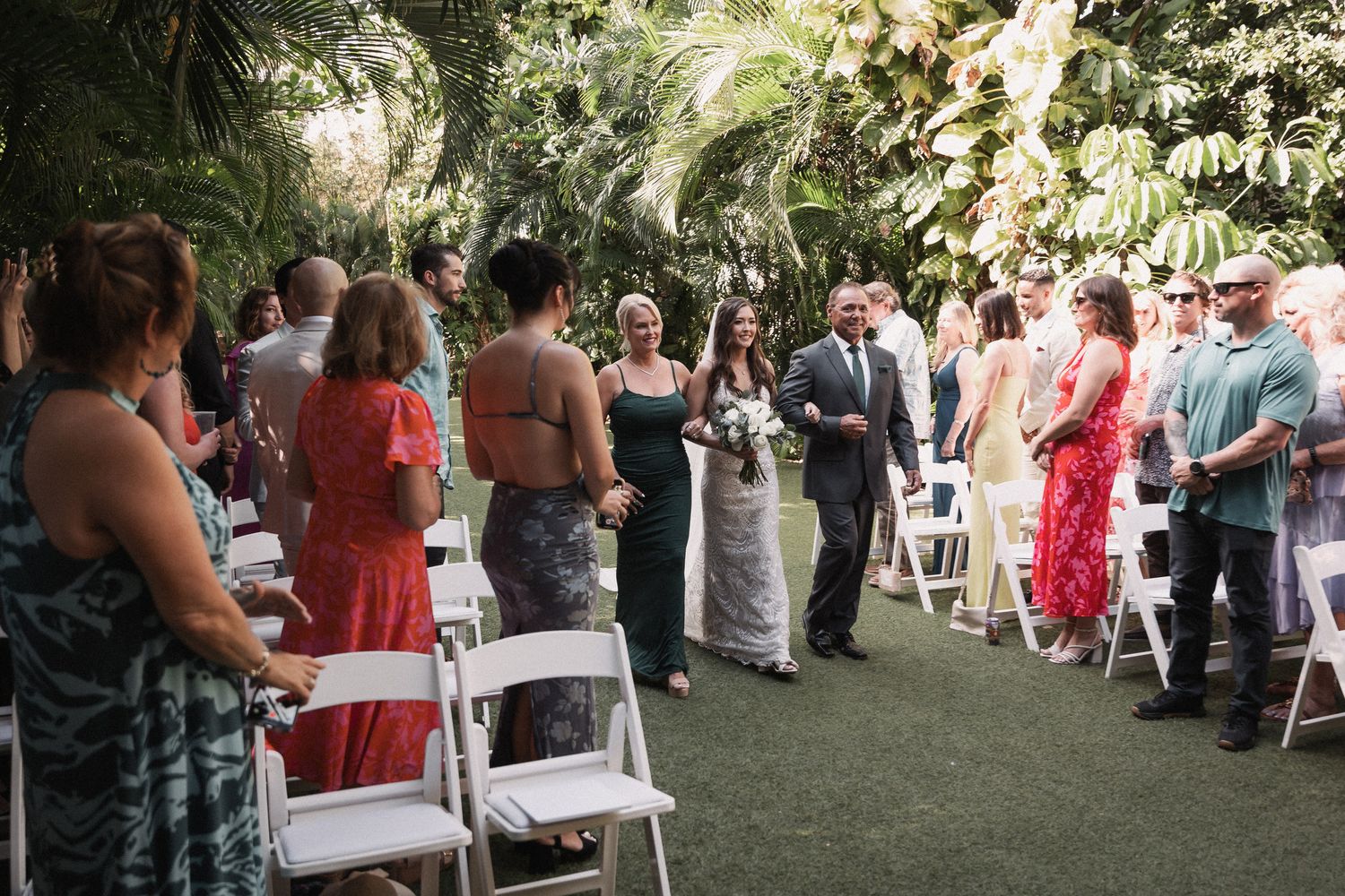 Wedding processional outdoors with guests seated in white chairs amid tropical greenery.