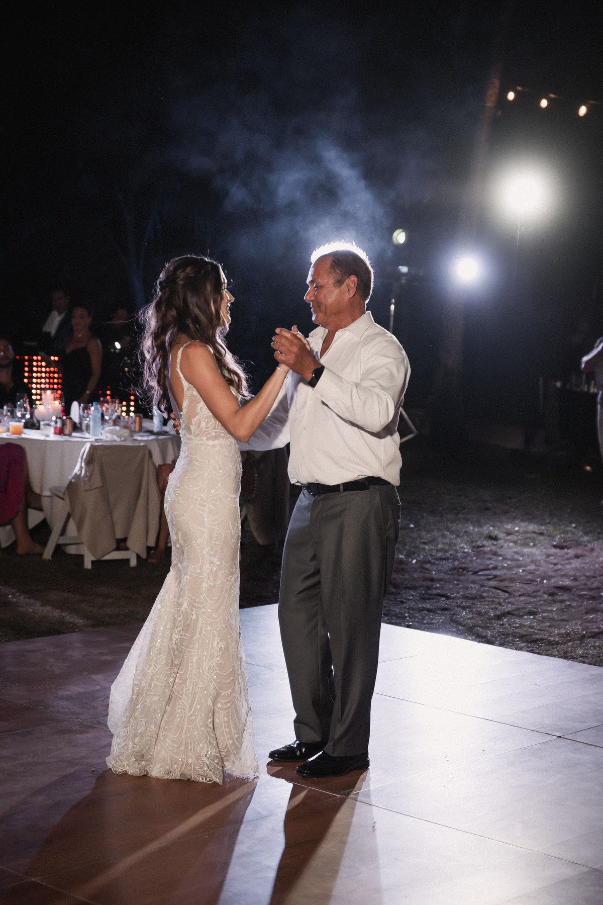 Bride in lace gown shares emotional father-daughter dance on lit outdoor dance floor.