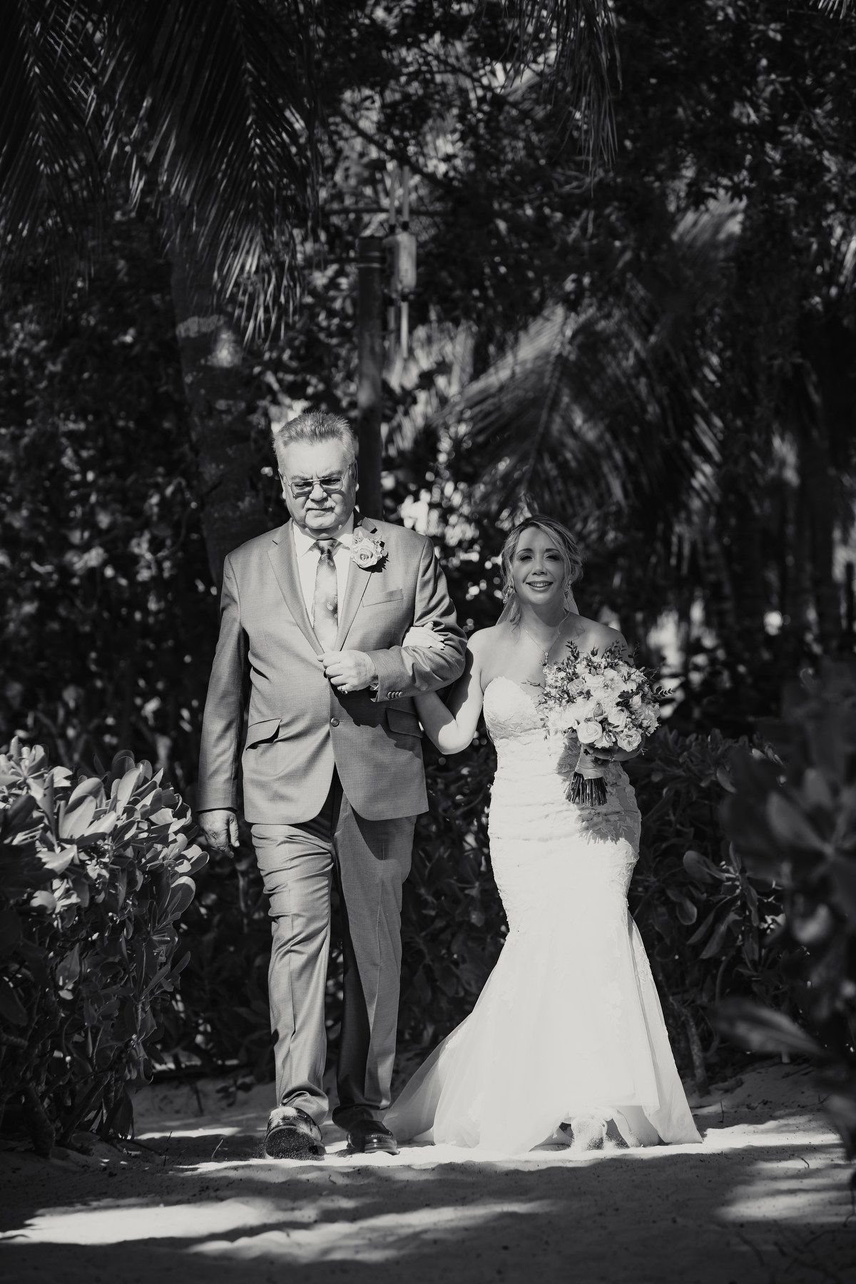 Black and white photo of bride walking down aisle with father in tropical garden setting.