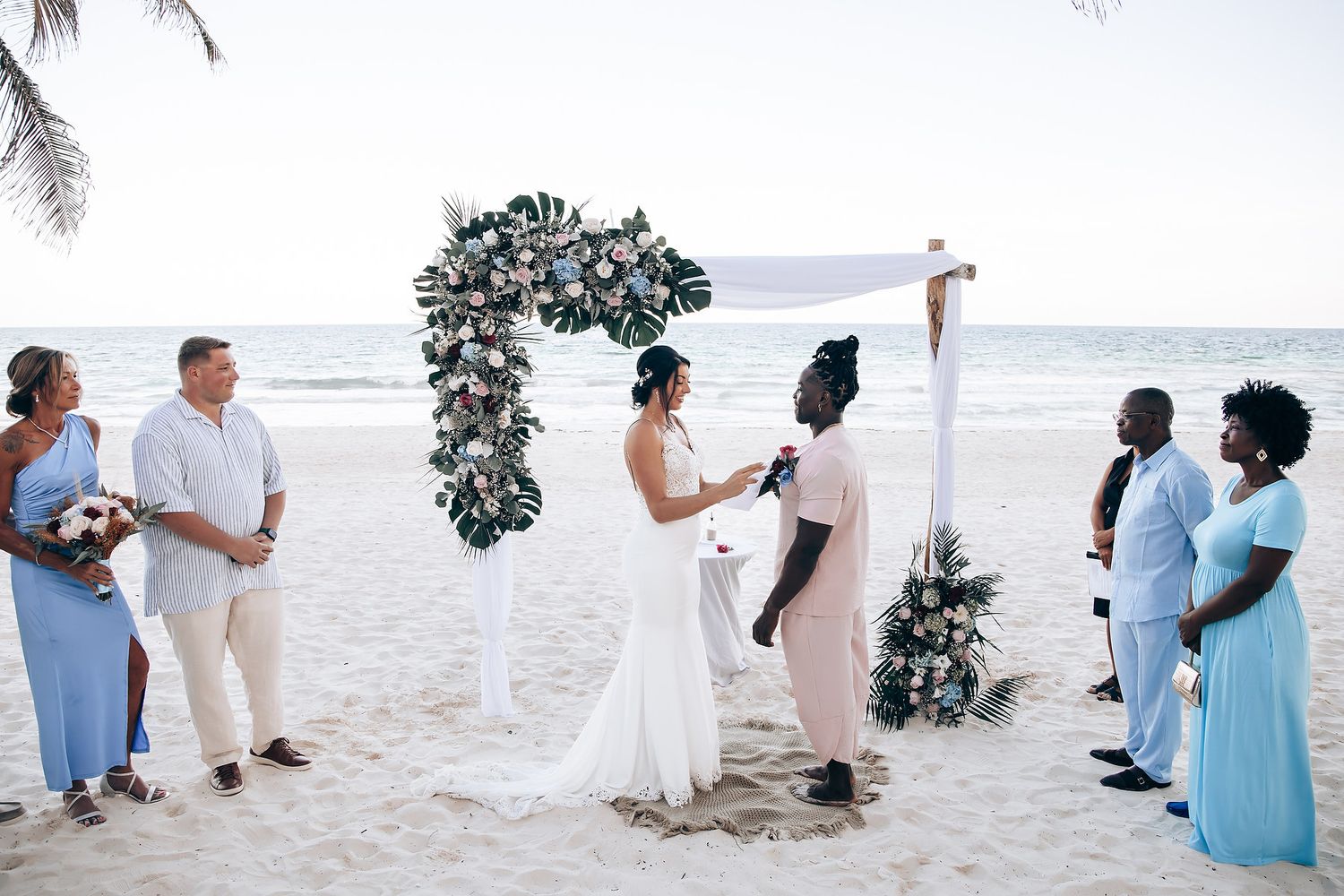 A couple exchanges vows on a beach, surrounded by guests and a floral arch.