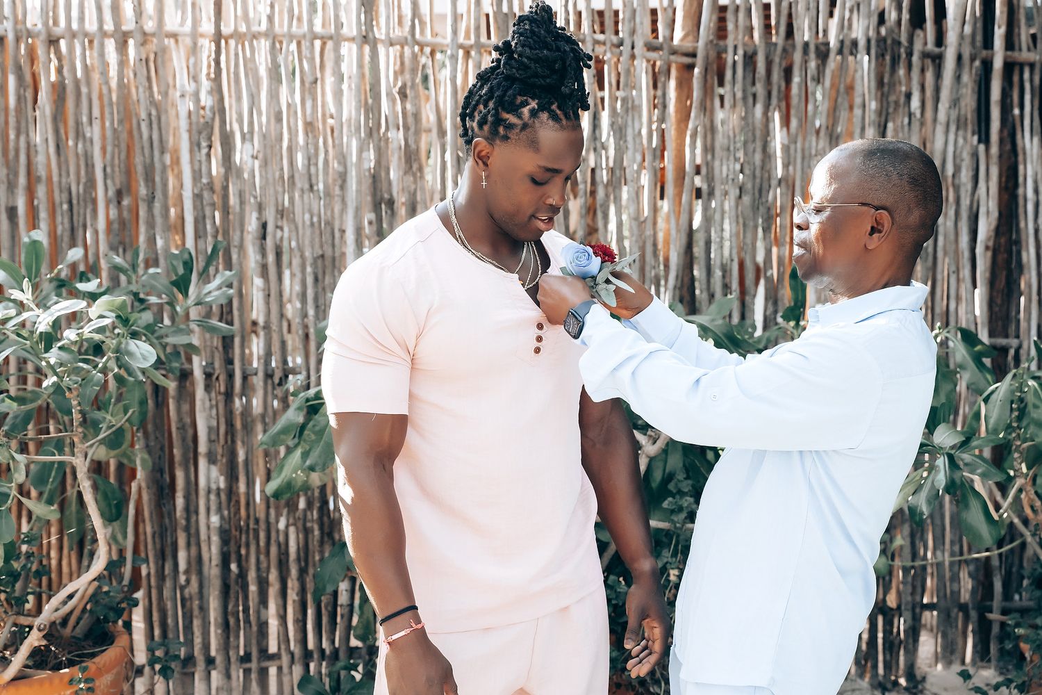 Two people, one in a white outfit and the other in a pink outfit, are standing together in front of a bamboo fence.