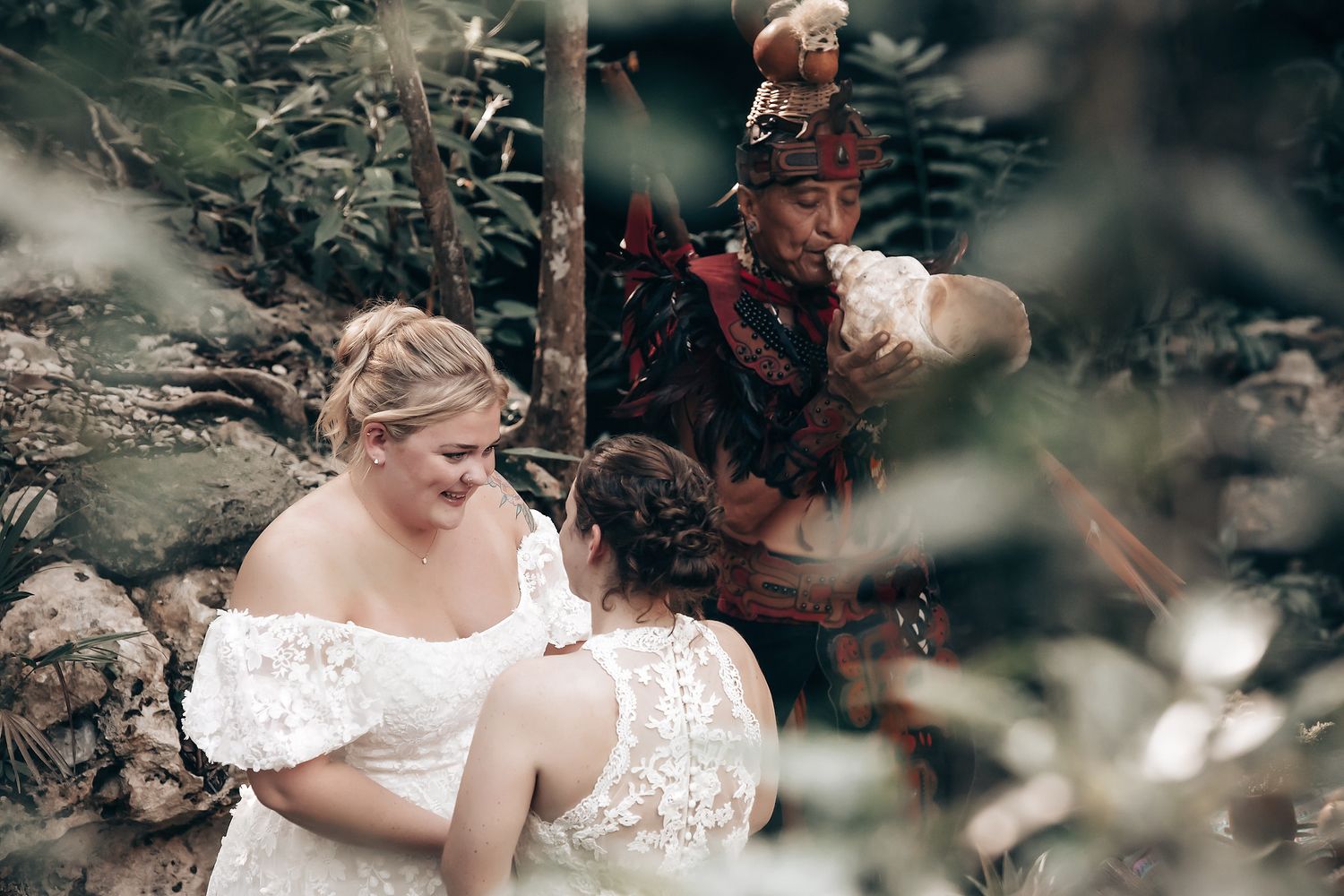 Two women in white wedding dresses standing in a lush, forested setting.