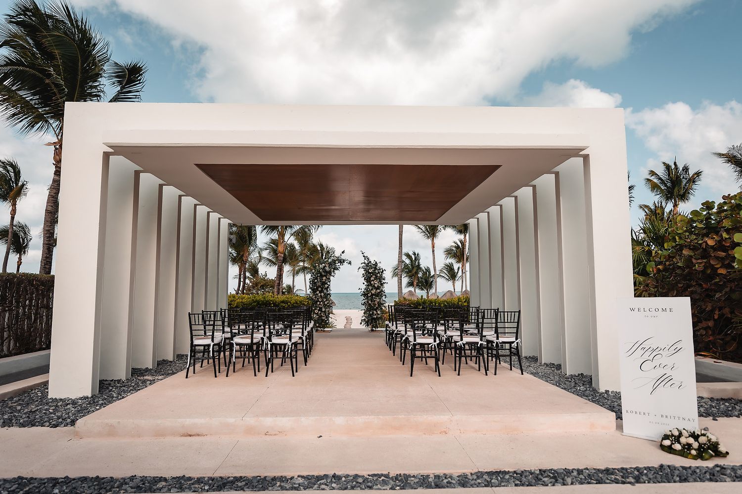 Elegant outdoor wedding ceremony setup with black chairs under a modern white pergola near the ocean.