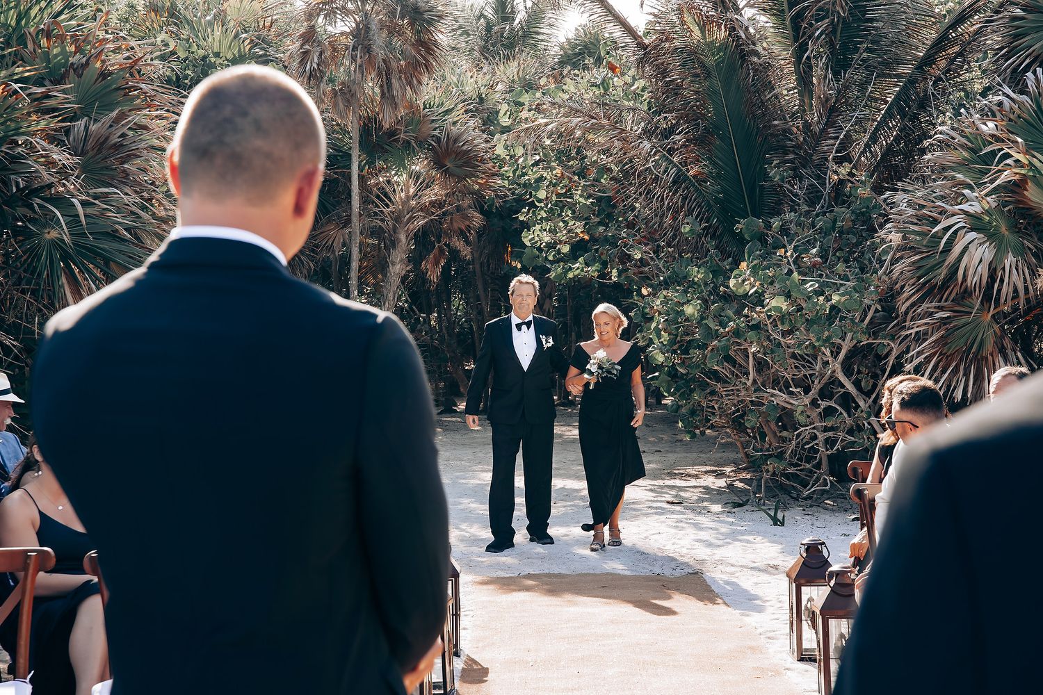 An outdoor wedding ceremony takes place in a lush tropical garden setting with guests seated.