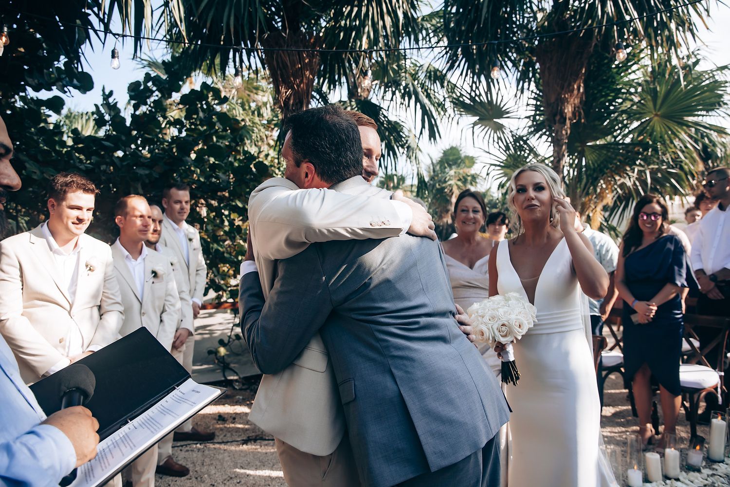 An emotional outdoor wedding ceremony moment under palm trees with guests looking on.