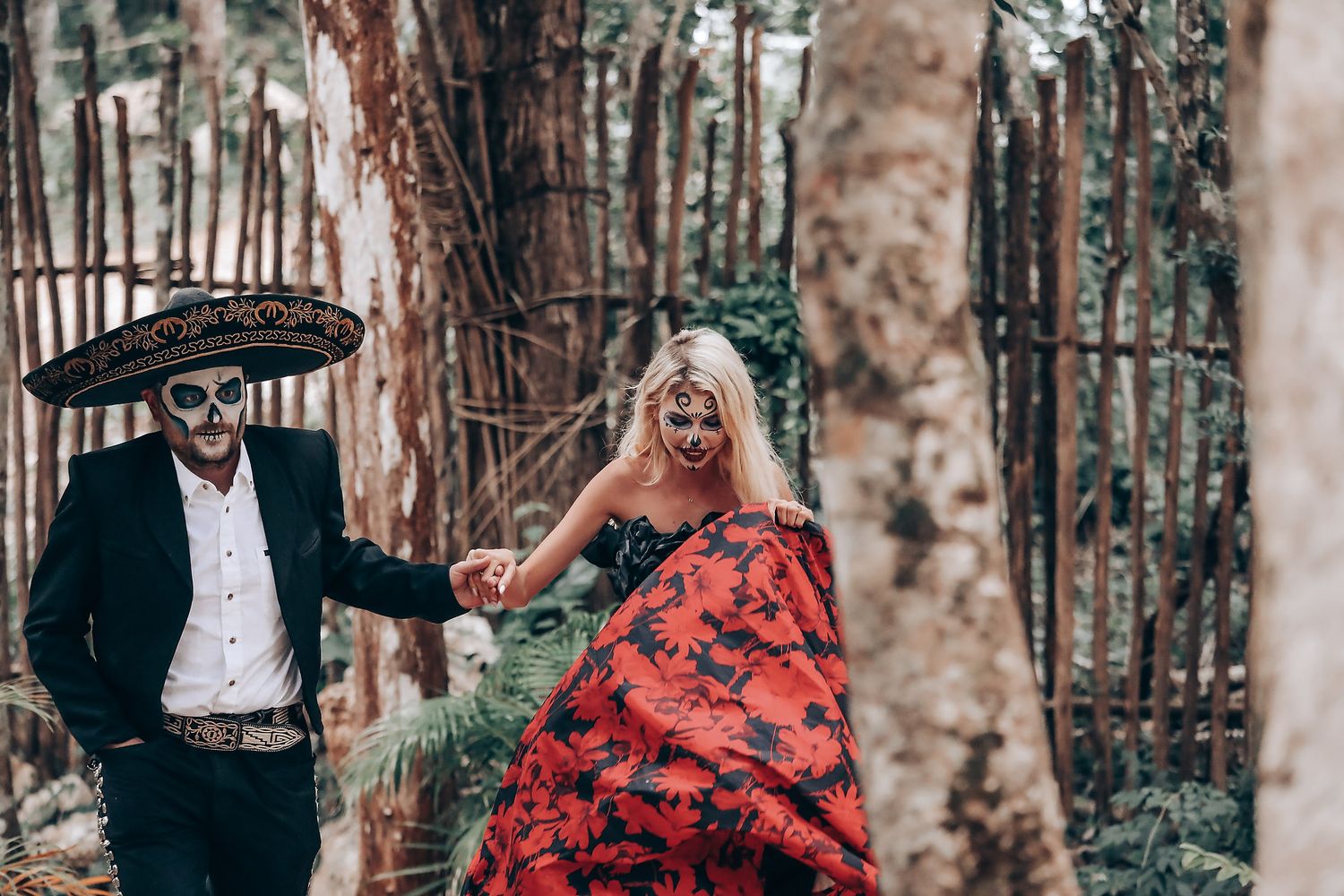 Couple in gothic wedding attire poses beneath trees with dramatic red dress details.