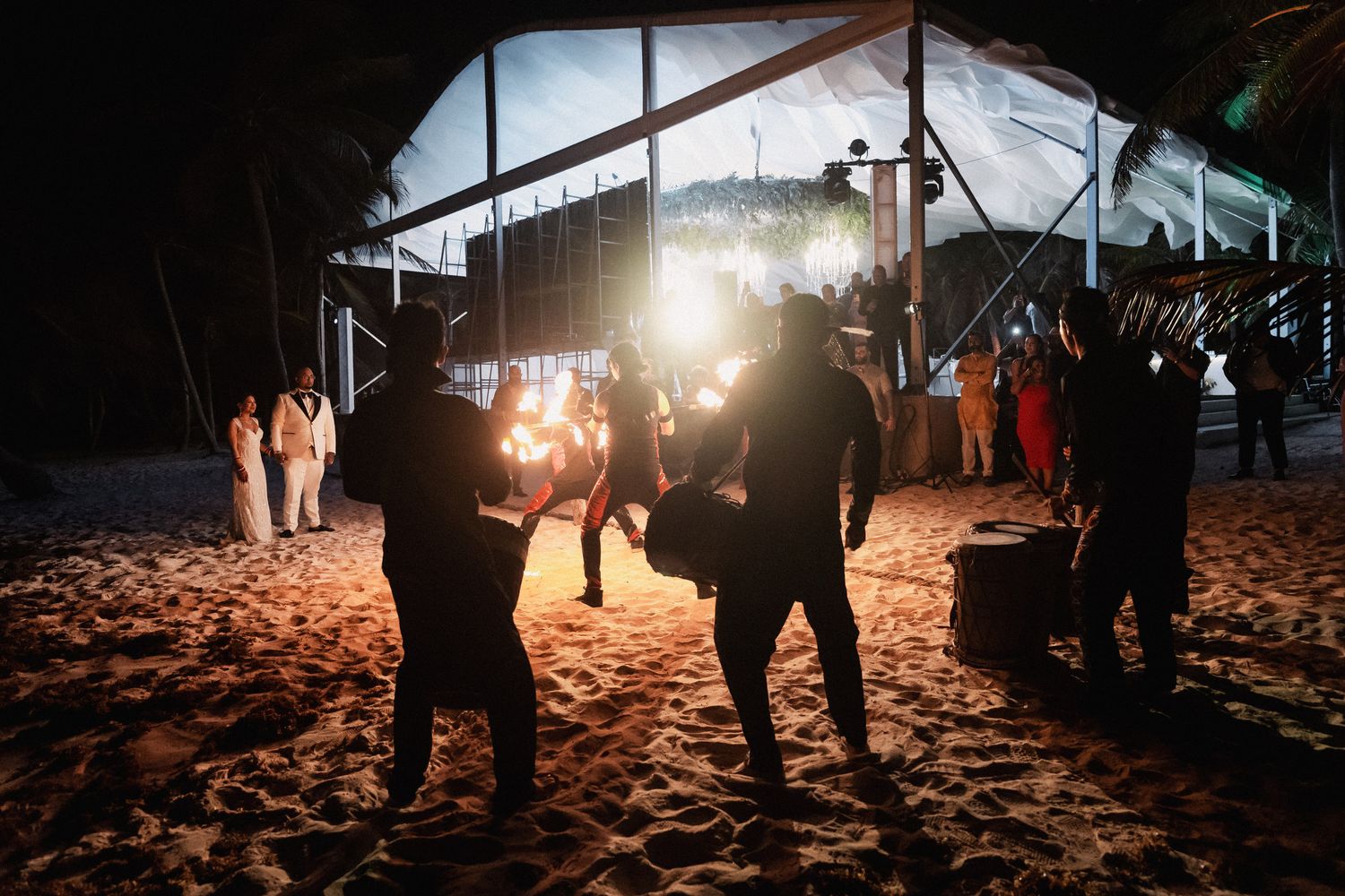 Musicians performing on sandy beach outside illuminated wedding reception tent at night.